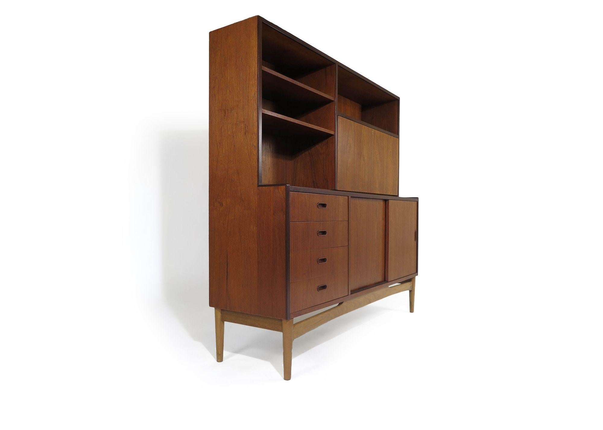 1965 Danish tall cabinet crafted of teak with sliding doors, series of four drawers, a locking drop-front desk surface, and open bookcase with adjustable shelves. The cabinet is raised on a sculptural solid oak base with tapered legs. The cabinet