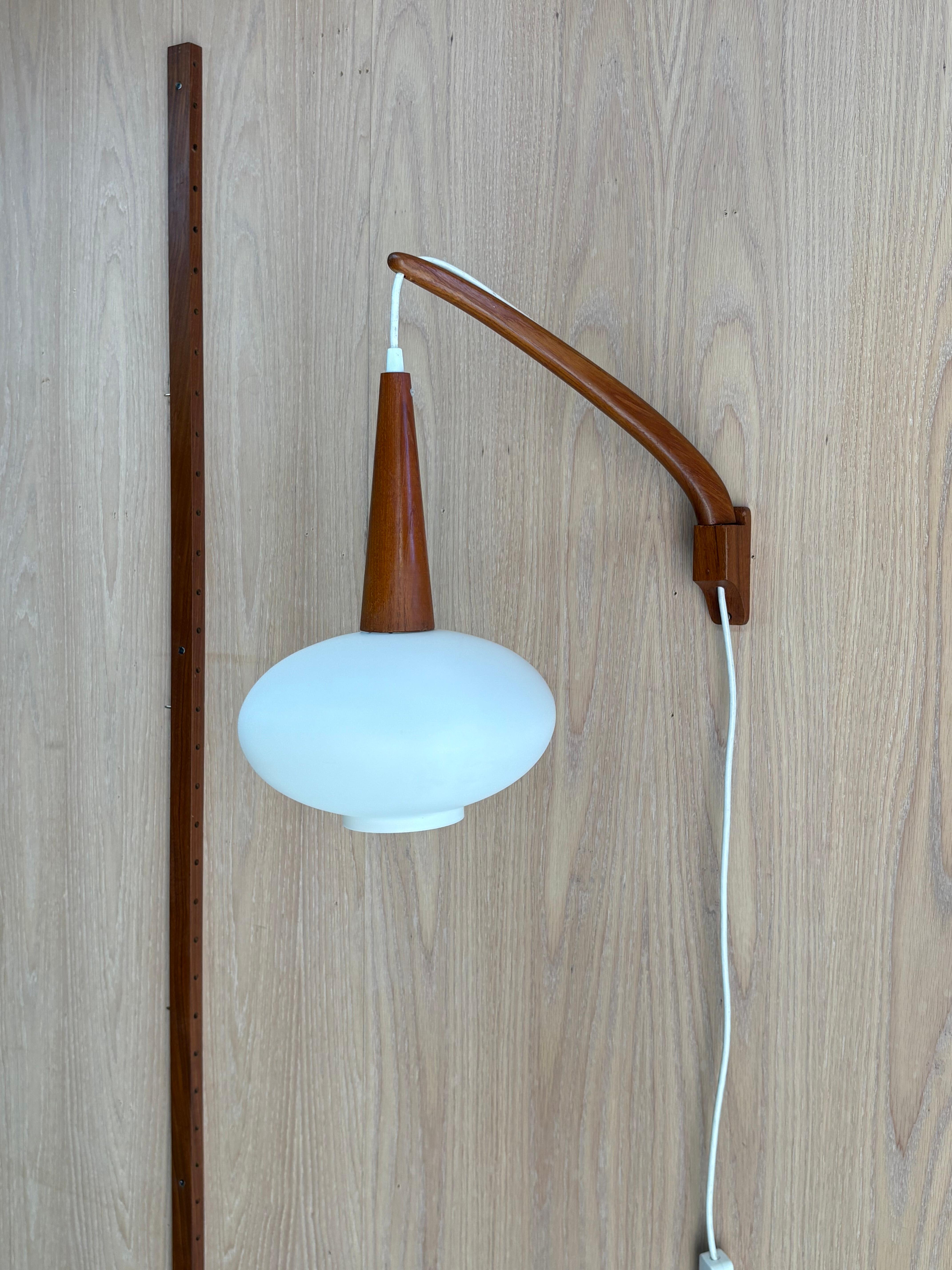 A striking mid-century teak wall light from the 1960s, blending warm organic materials with functional design. The sculpted teak arm pivots smoothly from side to side, allowing the light to be directed as needed, while the frosted glass shade casts