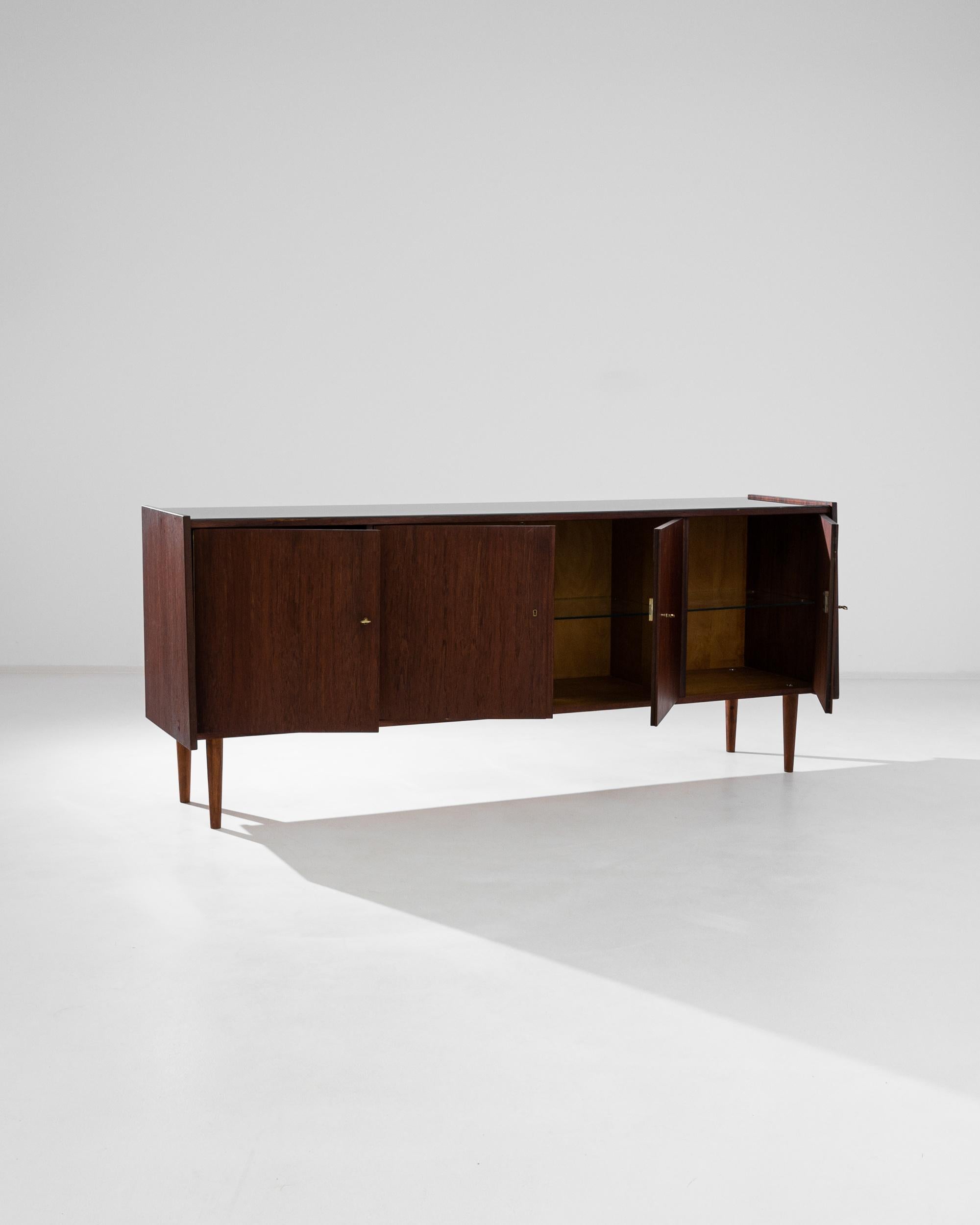 A wooden sideboard with glass top from Denmark, produced circa 1960. A cool, classic Mid-Century Modern sideboard constructed of dark teak laid in planes that create chic angles. This handsome piece features four locking cabinets with a glass shelf