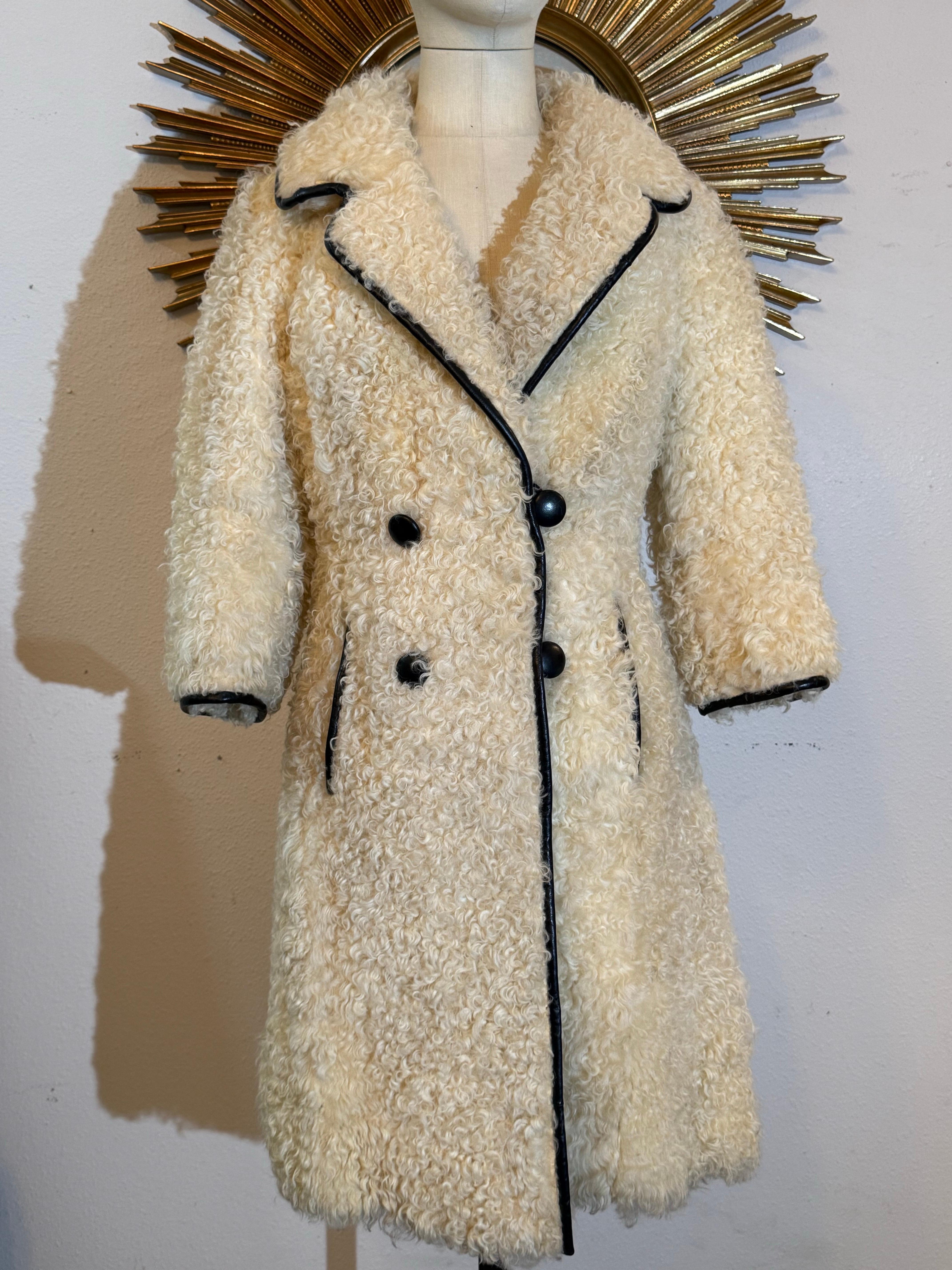 Cappotto in pelliccia d'agnello persiana mod. crema di Donald Brooks del 1960 con profili in pelle nera. Doppiopetto in stile A-line con cintura sulla schiena e tasche laterali a fessura. Collo profondo e dentellato che può essere ribaltato.