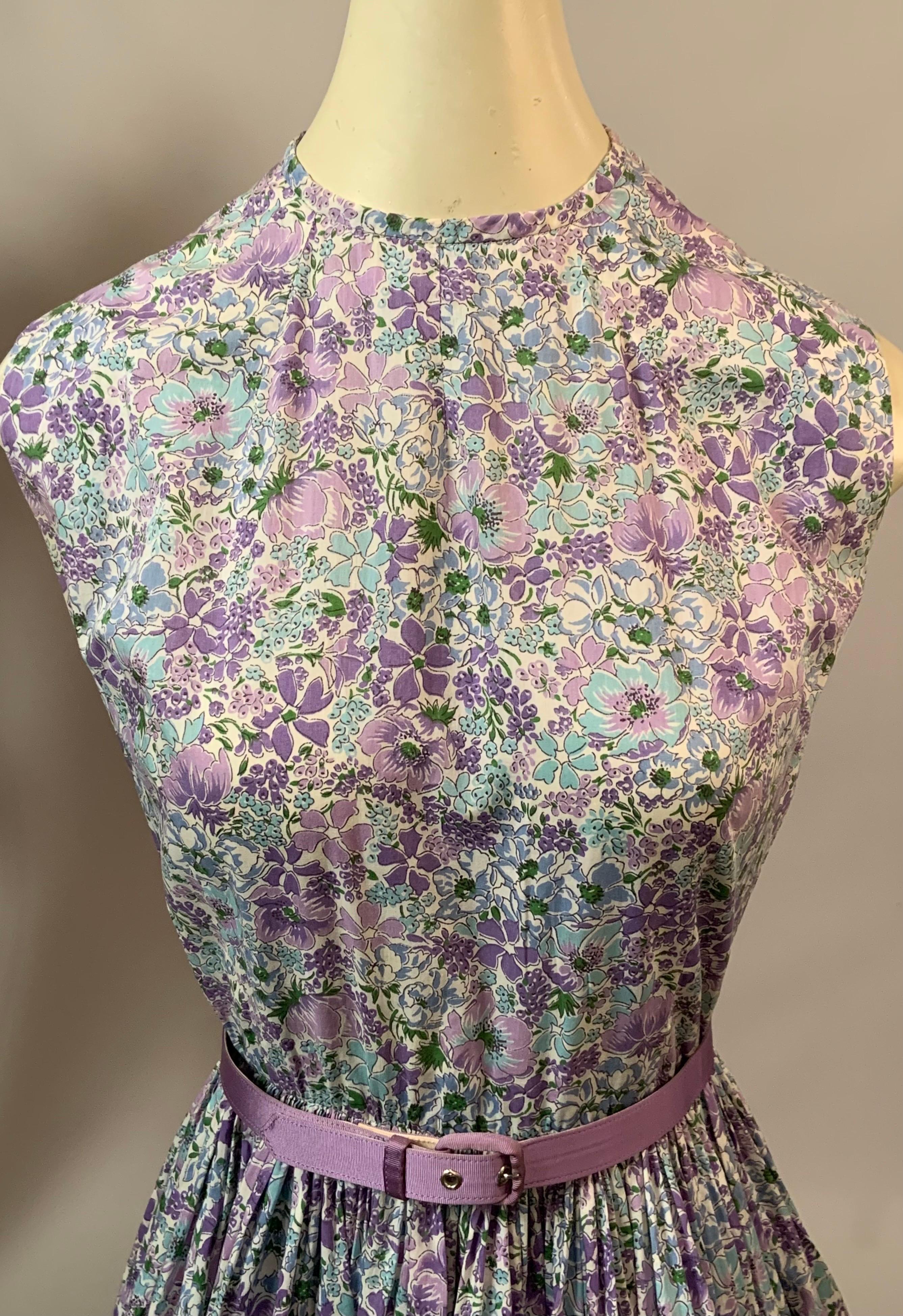 This charming dress is made with Liberty Of London fabric yardage in a palette of blues, lavenders, purple and green on a white background.  It is sleeveless with a rounded neckline, fitted waist and a full skirt.  There s a tie bow at the center