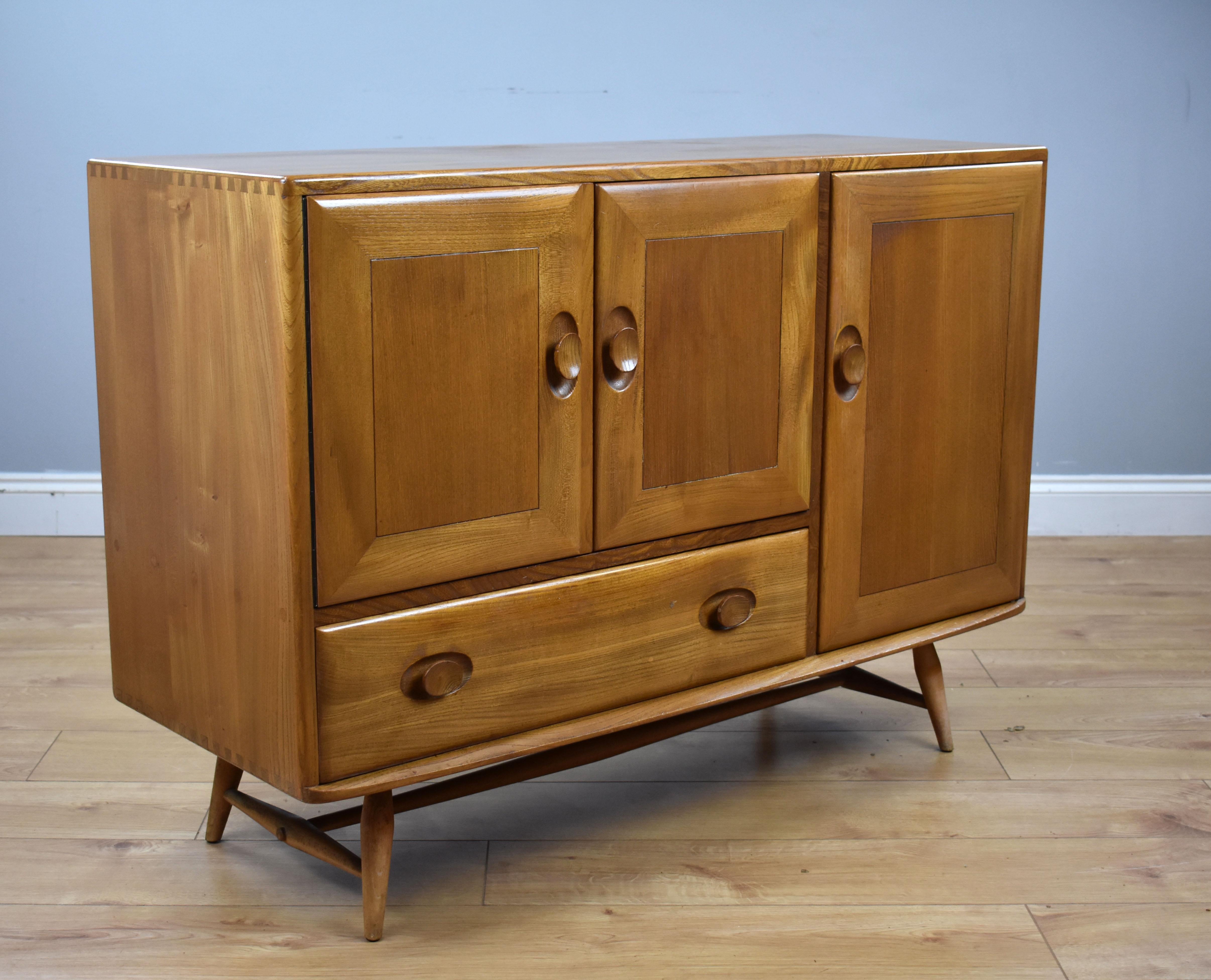 For sale is an Ercol elm Windsor sideboard, having a pair of cupboard doors opening to a single shelf, below this a single drawer. The right hand side there is another cupboard door opening to a fitted cutler drawer to the top above a shelf below.