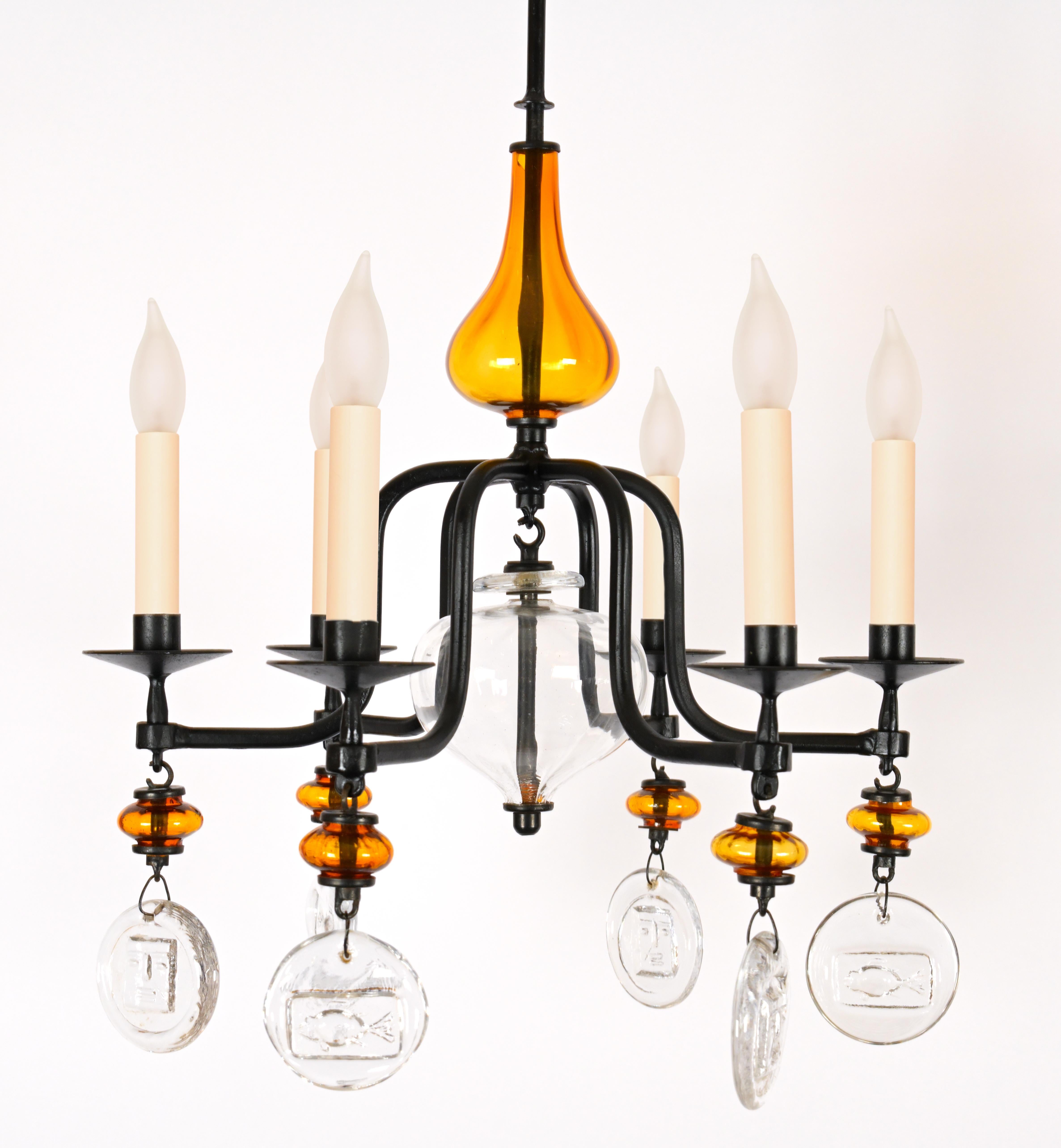 A glass and iron chandelier by Swedish Designer Erik Hoglund (1932-1998) having a bulbous orange blown glass center with 6 curved arms decorated with circular glass chips with imprints of playful faces and symbols. Includes standard wiring.