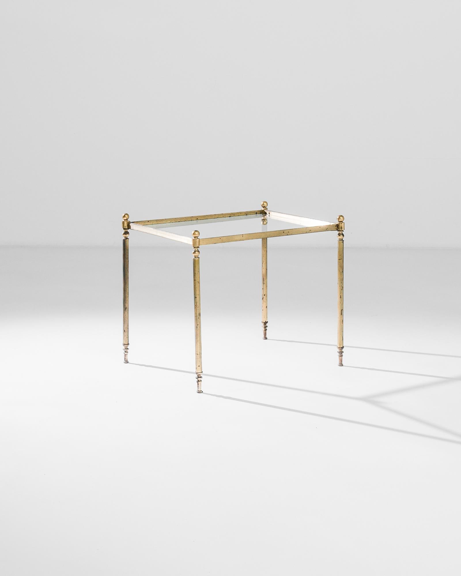 Light footed yet lavish, this brass coffee table from 1960s France has an Art Deco air of sophistication. A square tabletop of clear glass is set within a frame of fluted brass. The slender pillars of the legs are crowned by metal orbs; tapered feet