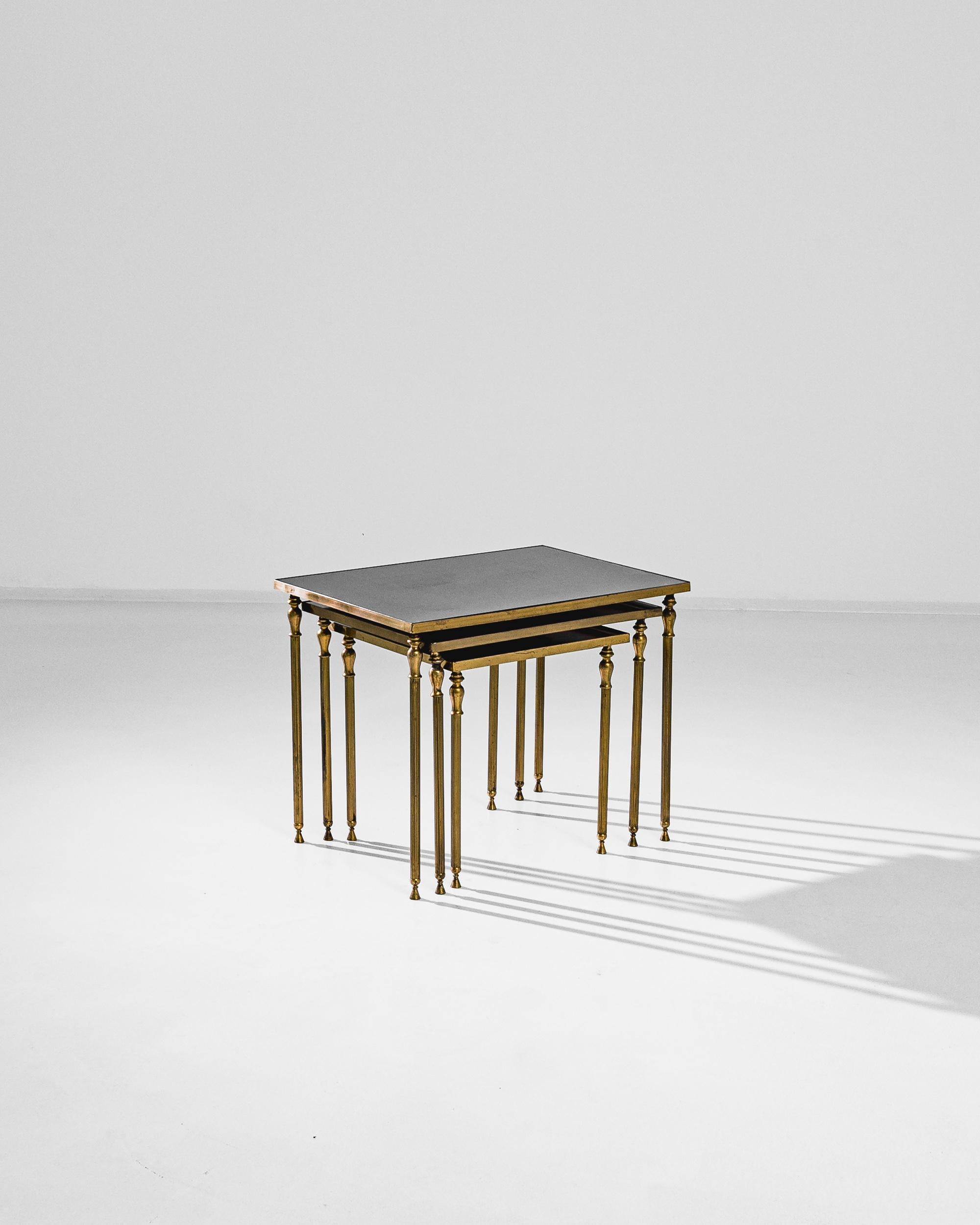 This trio of nesting tables provides a sophisticated accent. Made in France in the 1960s, a gilded brass frame supports a tabletop of obscure, smoke-grey glass. Fluted legs, crowned with golden urns, create a Neo-classical silhouette — while the