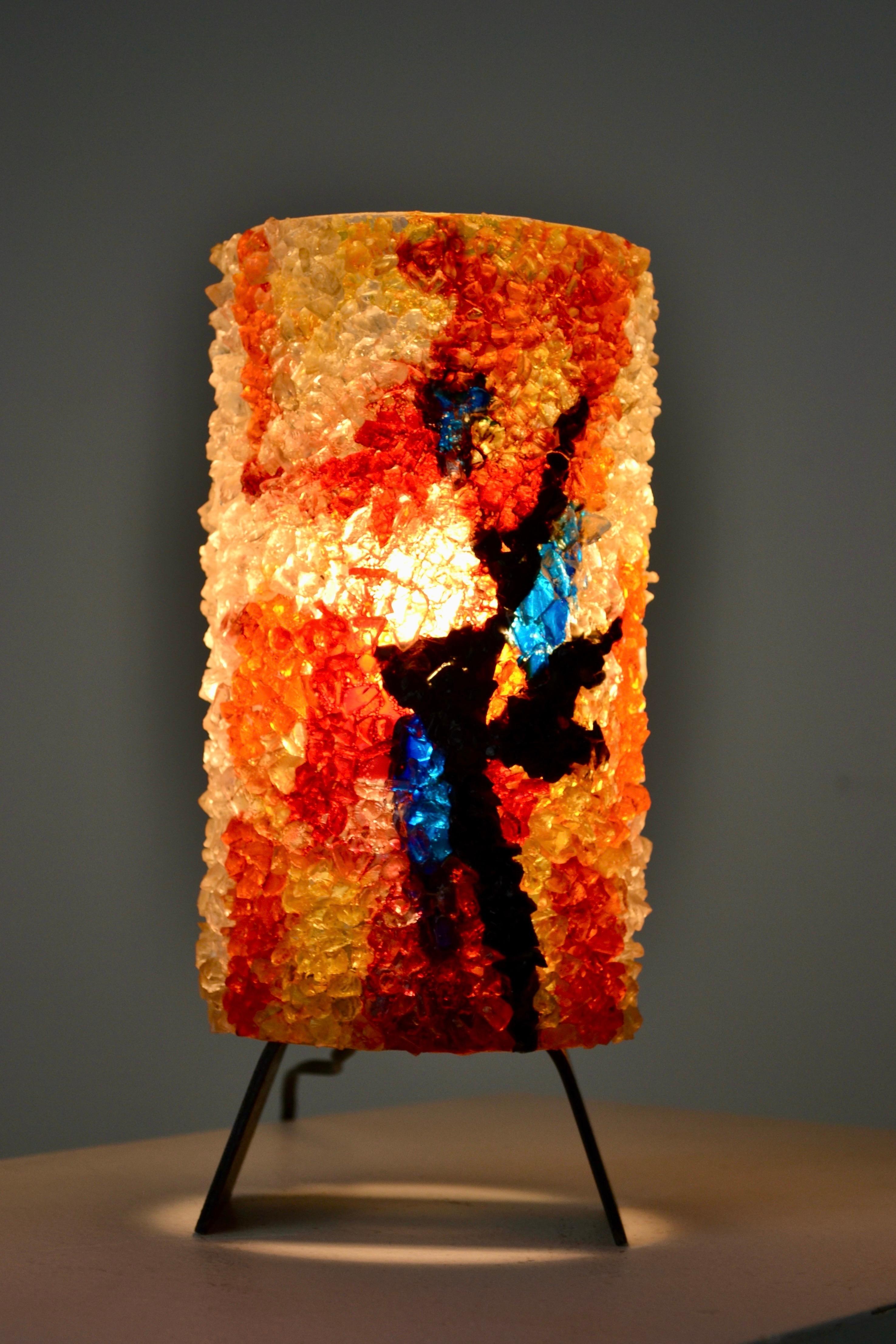 À markante Tischleuchte aus Frankreich aus den 1960er Jahren. Der dreibeinige Sockel ist aus Stahl gefertigt und trägt einen zylindrischen Diffusor aus strukturiertem Kunststoff mit lebhaften Einschlüssen aus farbigem Harz in warmen Tönen, der bei
