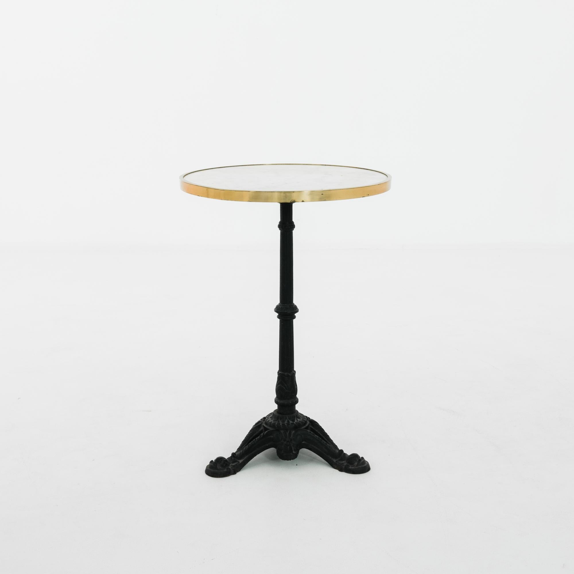 A cast-iron table from 1960s France with an elegant bistro aesthetic. The profile of the cast iron column, set atop embellished tripod feet, creates a graceful support for a round marble tabletop, ringed with brass. The subtle grey veining of the