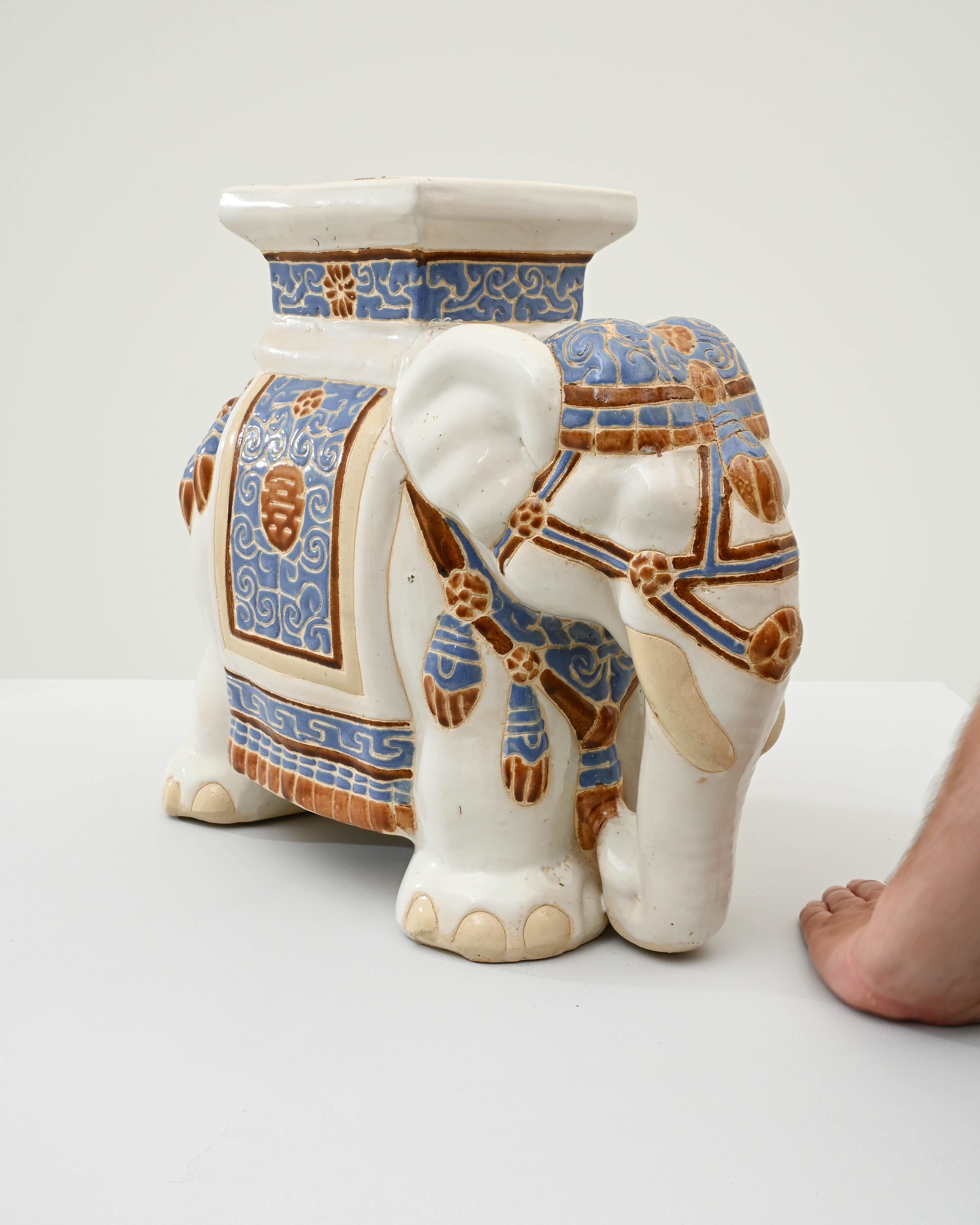 A ceramic decoration from 1960s France in the shape of an elephant. A saddle seat and blanket are glazed with pale blue and ochre, the elephant’s skin painted with glossy white and laced with lines of the earth tone clay body; the assortment of