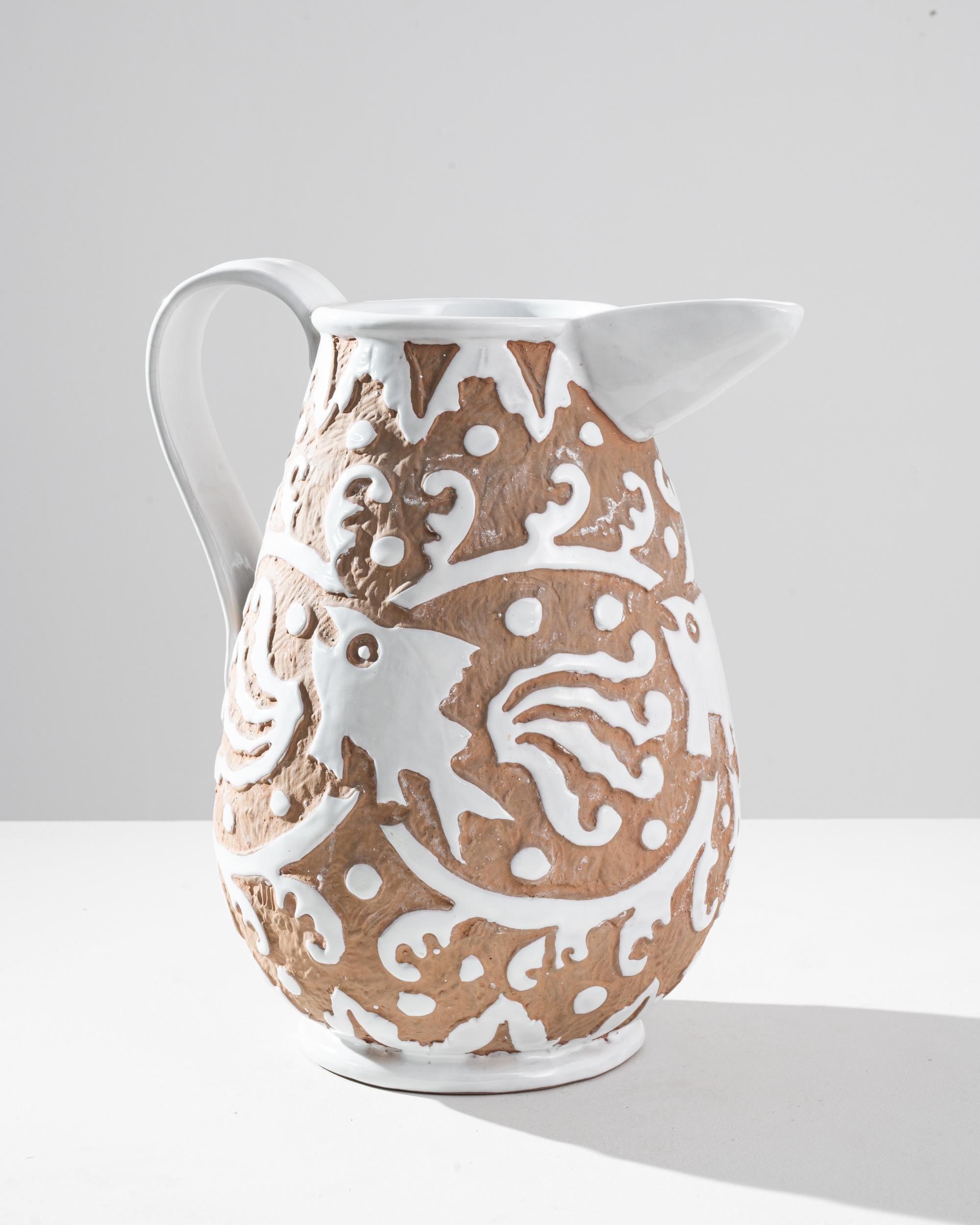 This vintage ceramic jug combines rustic simplicity with decorative charm. Made in France in the 1960s, an intricate country design stands out in white relief against a textural, terra-cotta colored background: it depicts a stylized fish, leaping