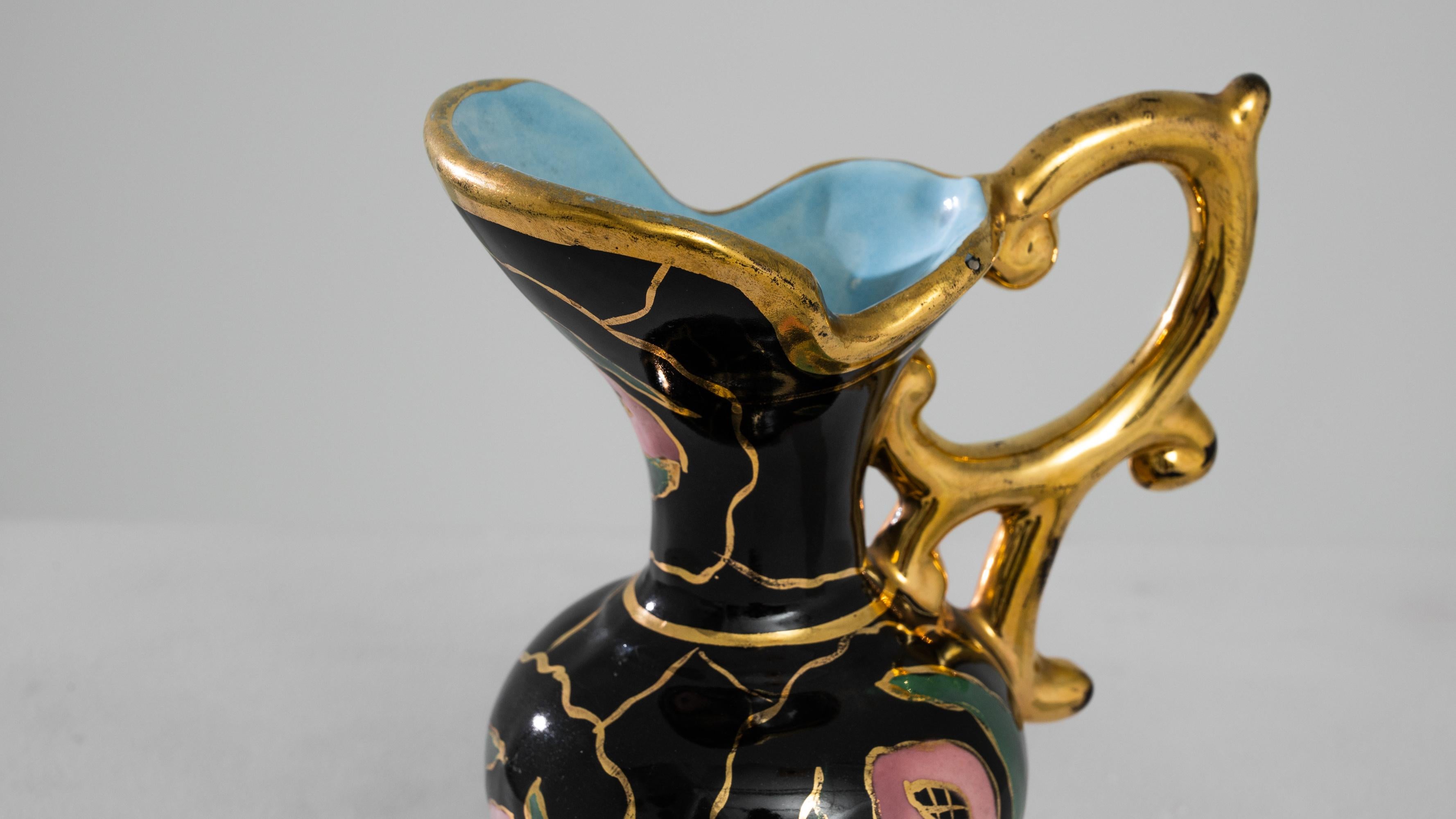 Behold the allure of this 1960s French ceramic jug, a paragon of elegance. Cloaked in a main black hue, it is adorned with delicate splashes of green and pink, reminiscent of blooming flowers. The intricate gold accents bestow a regal touch,
