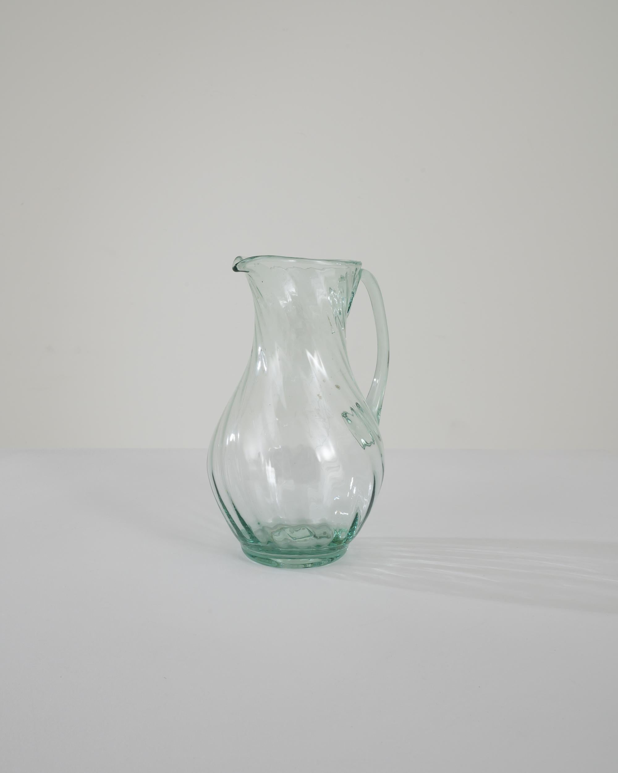 The simple grace of this vintage glass jug has a timeless charm. Hand-made in France in the 1960s, the fluid shape of the container is accentuated by the subtle sea-green hue of the transparent glass. Swirling ridges spiral up the glass, like water