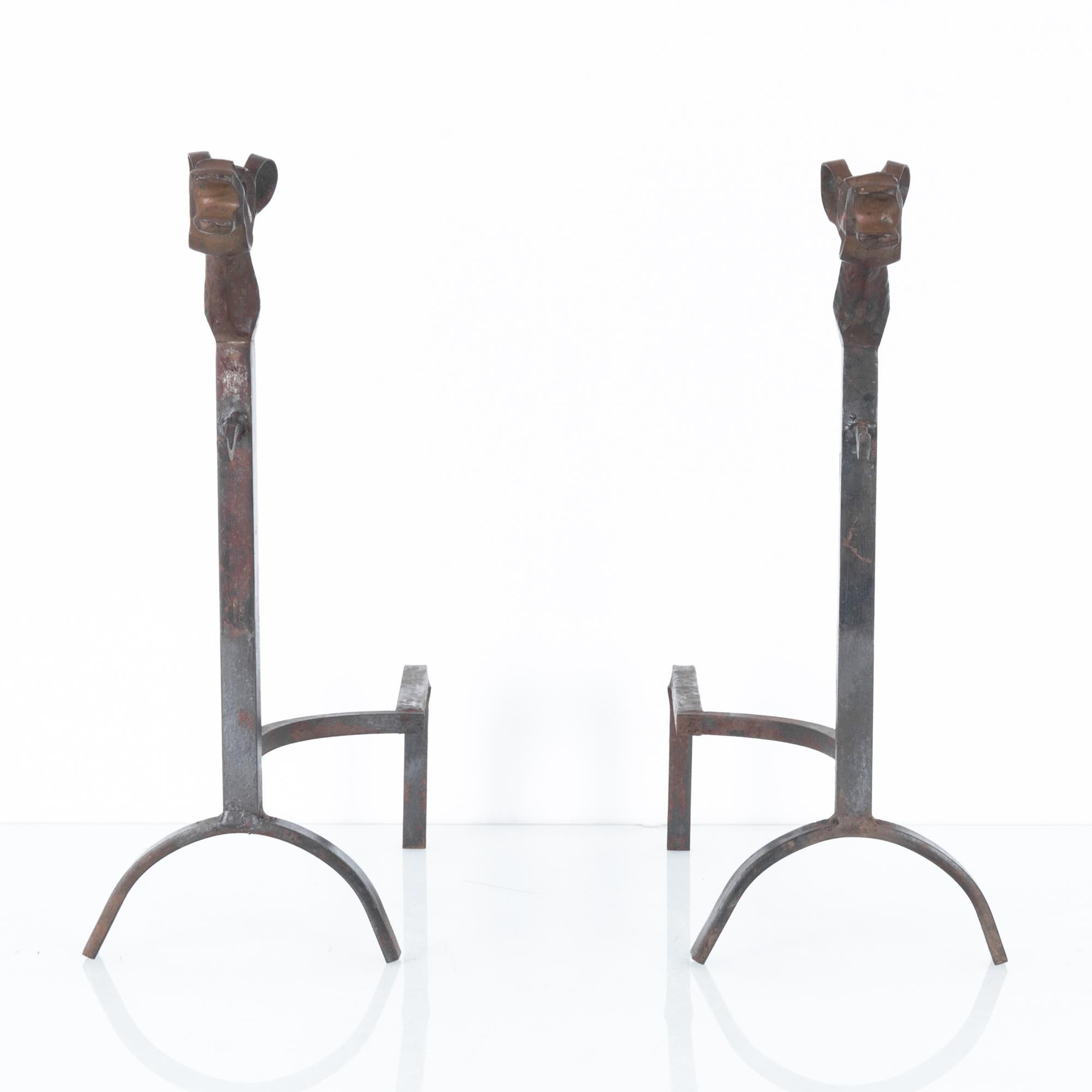 A pair of iron fire dogs from France, circa 1960. Two wrought iron brackets topped with open-mouthed iron animal heads. Originally used for burning logs in an open fire place, one bracket has a metal loop intended to support a poker and tongs. Small