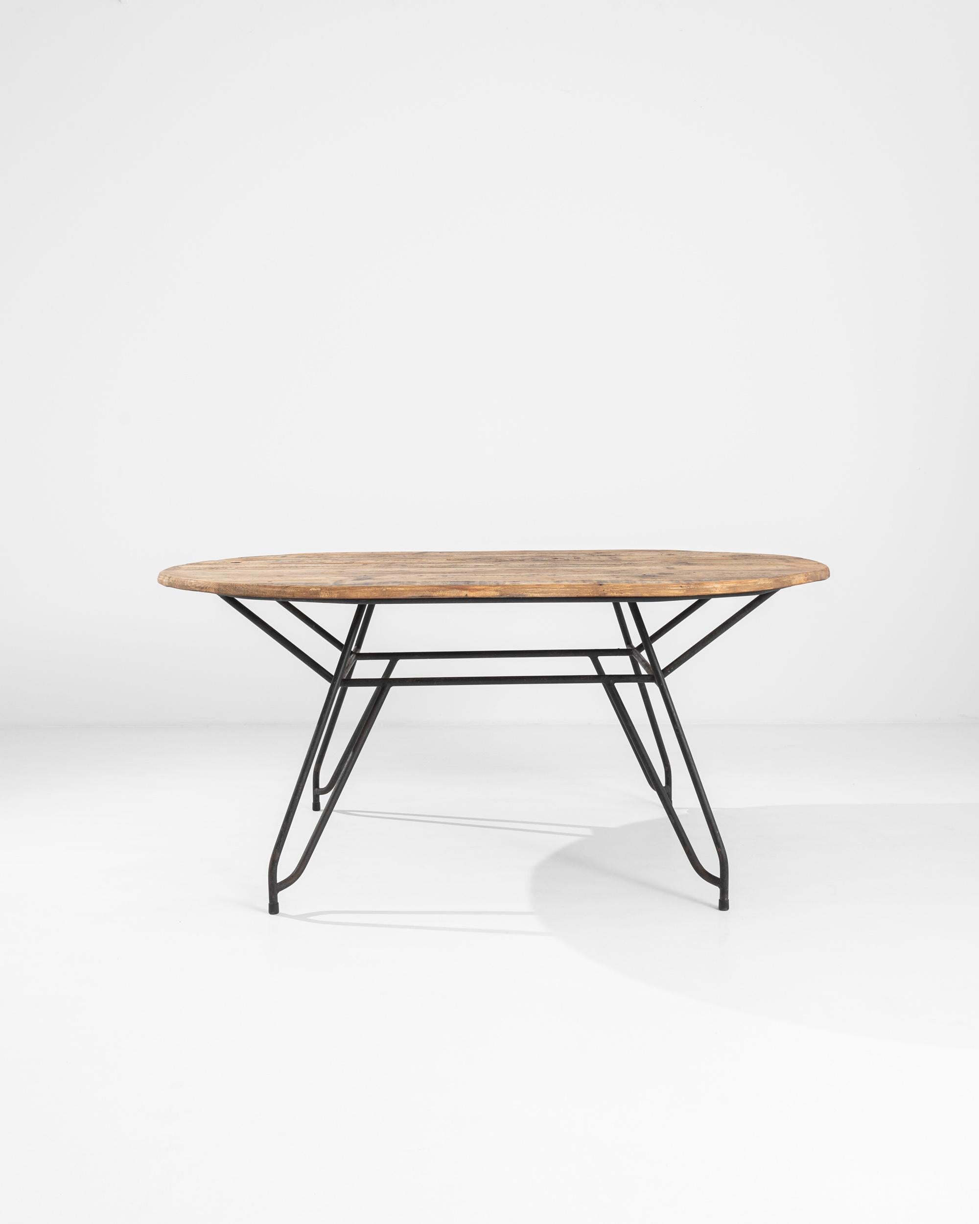 A metal midcentury French table with a wooden top. Solid hairpin legs extend diagonally at each corner, conjoined with a pleasing geometric network of supports. Solid planks of pleasantly worn lumber connect to form a charmingly rustic table top.