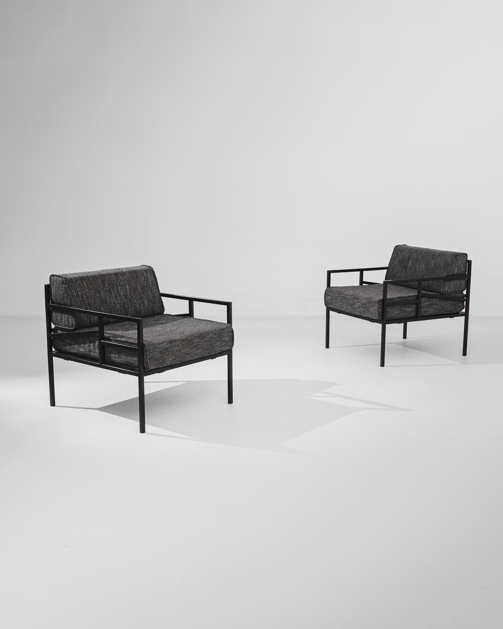 A pair of armchairs from 1960s France with a sleek Modernist aesthetic. A stylish, grid-like frame of black metal supports generous cushions upholstered in grey twill. Dynamic proportions and angular lines create an innovative contemporary