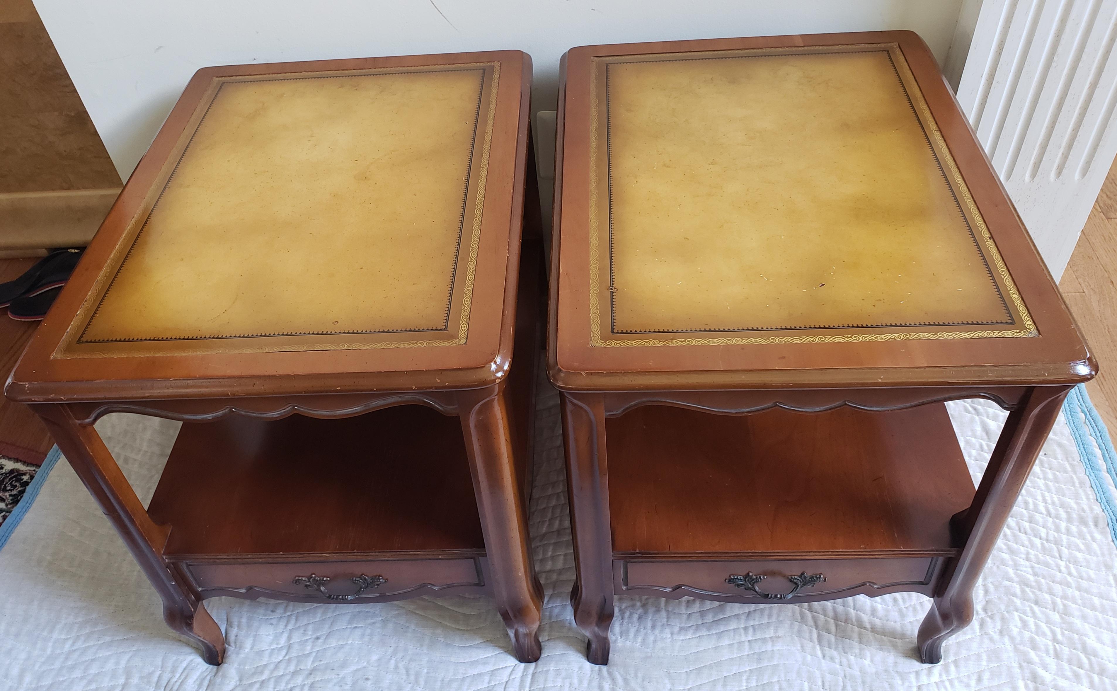 A pair of French Provincial style vintage side tables with leaves and Stinciling top. The tables feature a dark oak frame with a leather top insert, scalloped edges. Each table has a lower drawer with a scrolled pull and decorative cabriole legs.