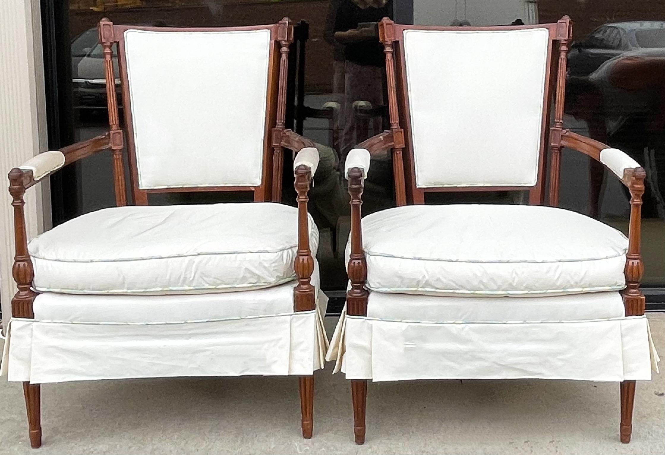 Too cute to leave behind! This is a pair of French style carved mahogany skirted bergere chairs. The attention to detail in the upholstery is not common any more. The ivory upholstery has plaid contrast welting. The arm covers could use replacing.