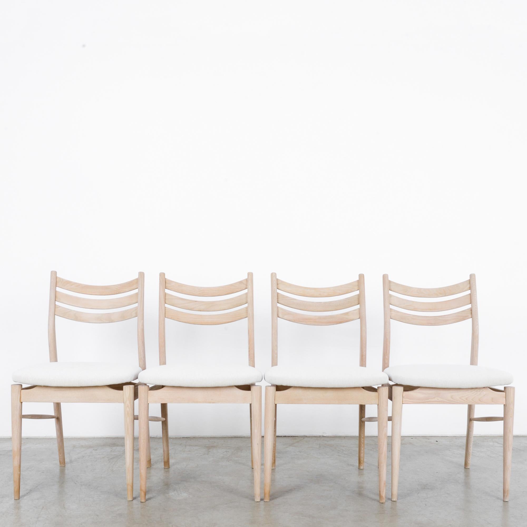 A set of four French wooden dining chairs, circa 1960s. Graceful legs connected by simple stretchers support a swooping apron holding aloft seats that appear as plump clouds, fog hugging the canopy of an old growth forest. With dramatic swooping