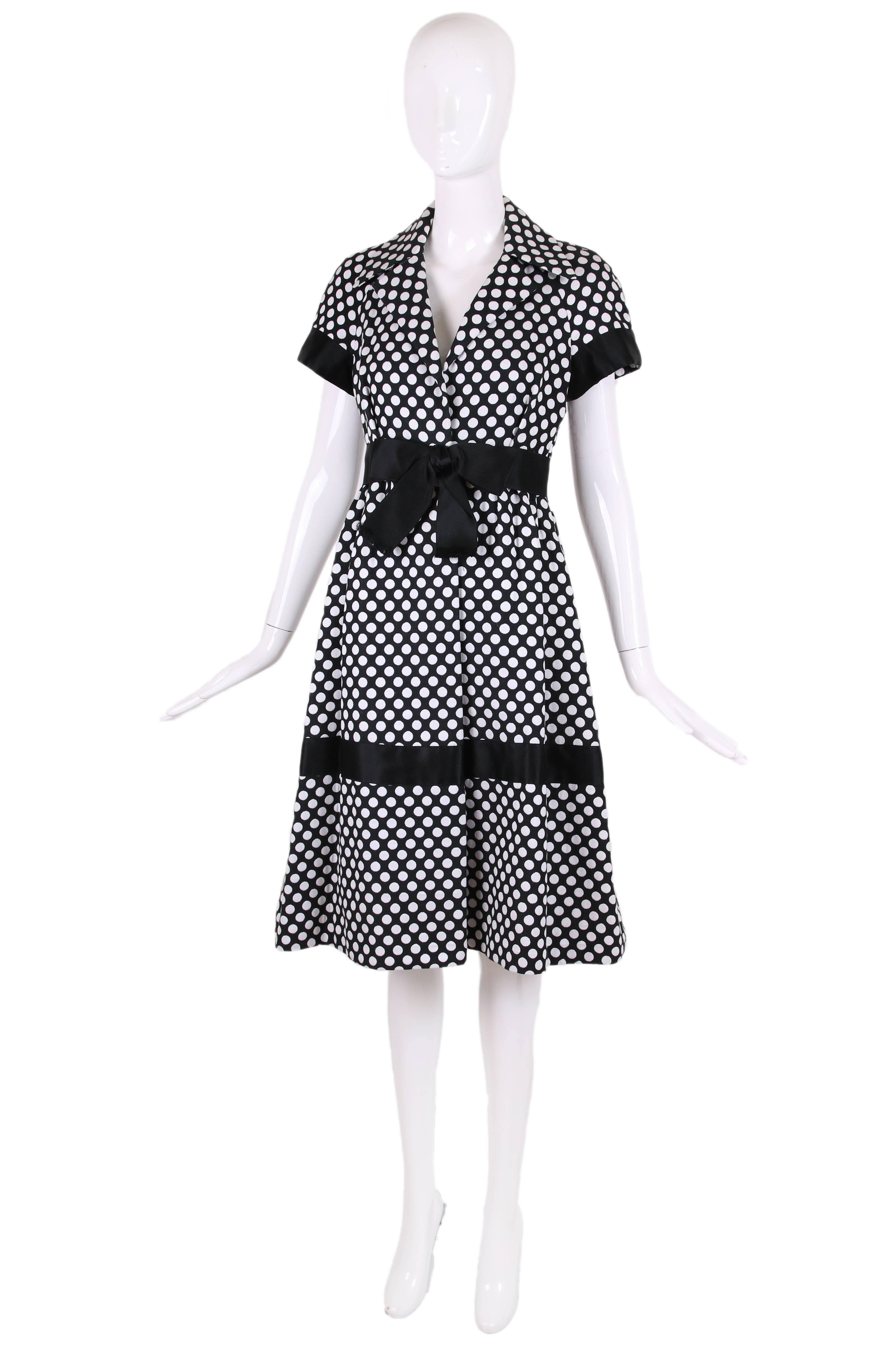 1960's Geoffrey Beene black and white polka dot dress with short sleeves, pleated flare skirt and black satin trim at sleeves, black satin bow at waist and a band of black satin trim mid-skirt. Hook and eye closure at interior waist. In excellent