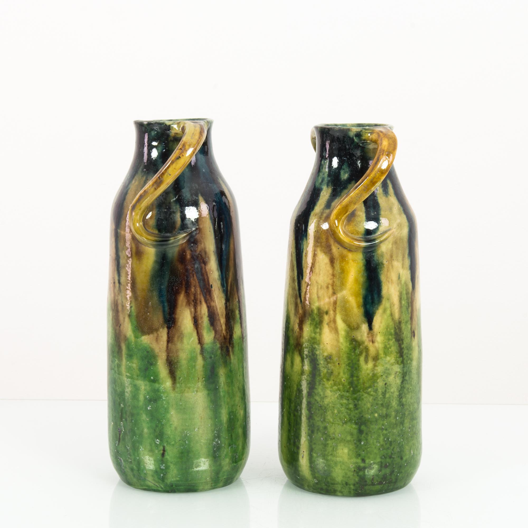 A pair of ceramic vases from West Germany, circa 1960. Streaks of forest colors create a dappled, sylvan effect — emerald, pine green, ochre. Twisting handles which emerge from the lip of the flasks add a whimsical touch. The subtle differences in