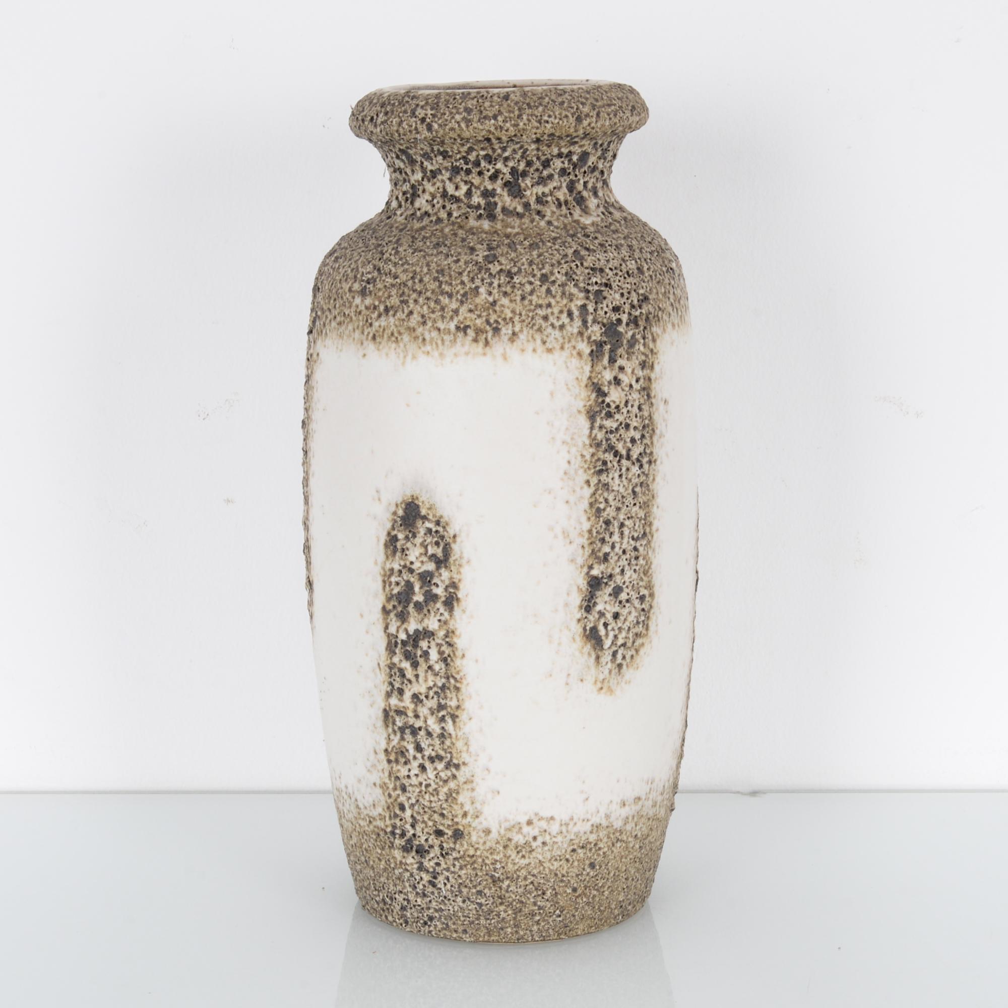 This ceramic vase was made in Germany, circa 1960. It has a textured, coral-like surface in shades of brown and gray, interrupted by ivory white swathes on the body. The rounded edges of its mouth, neck, and shoulders give the vase an elegant and