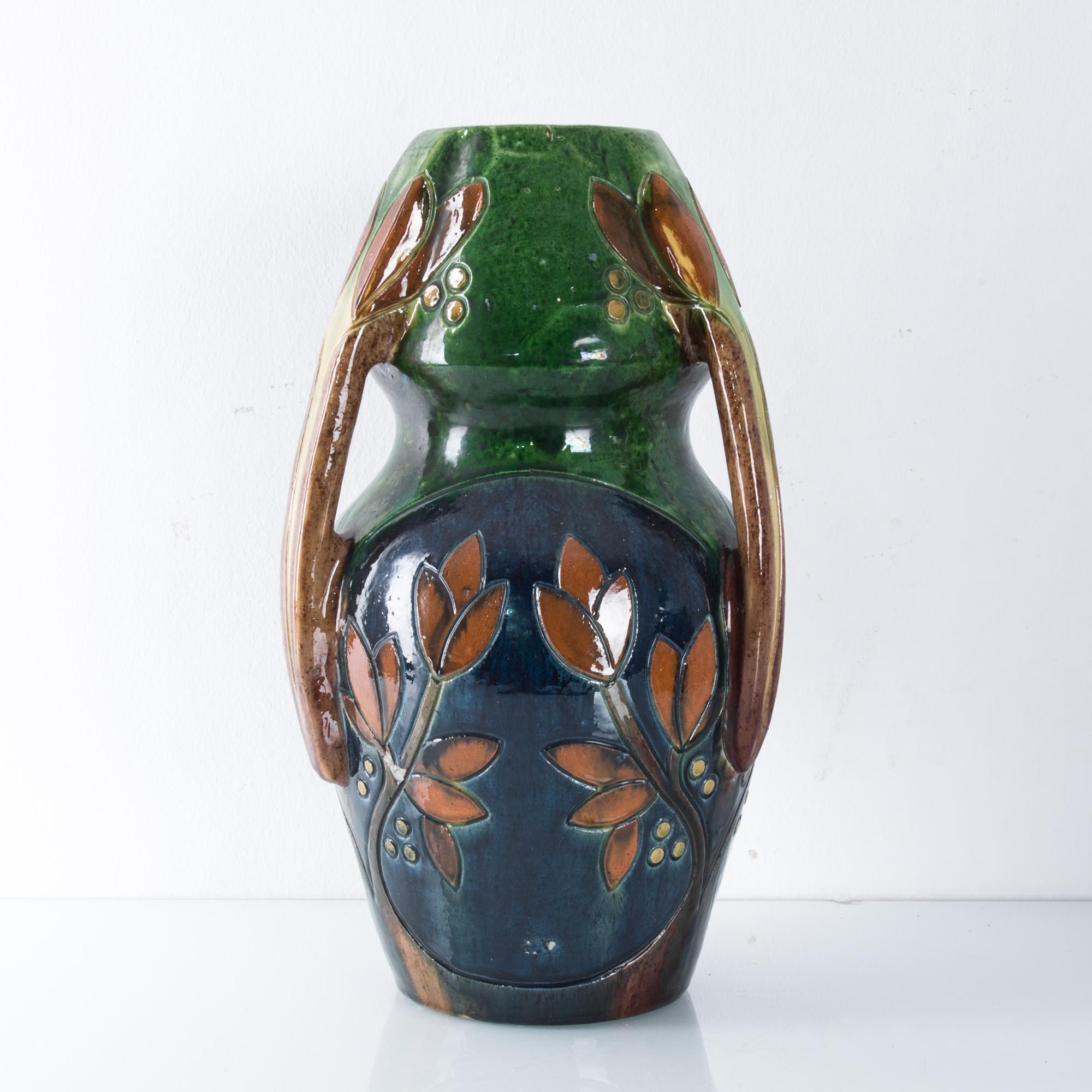 A ceramic vase from Germany, circa 1960. A leafy pattern is realized in a palette of cobalt blue, forest green and terracotta brown. The branches of the leaves extend to form handles, giving the vase a three-fold symmetry. The shape is wasp-waisted;