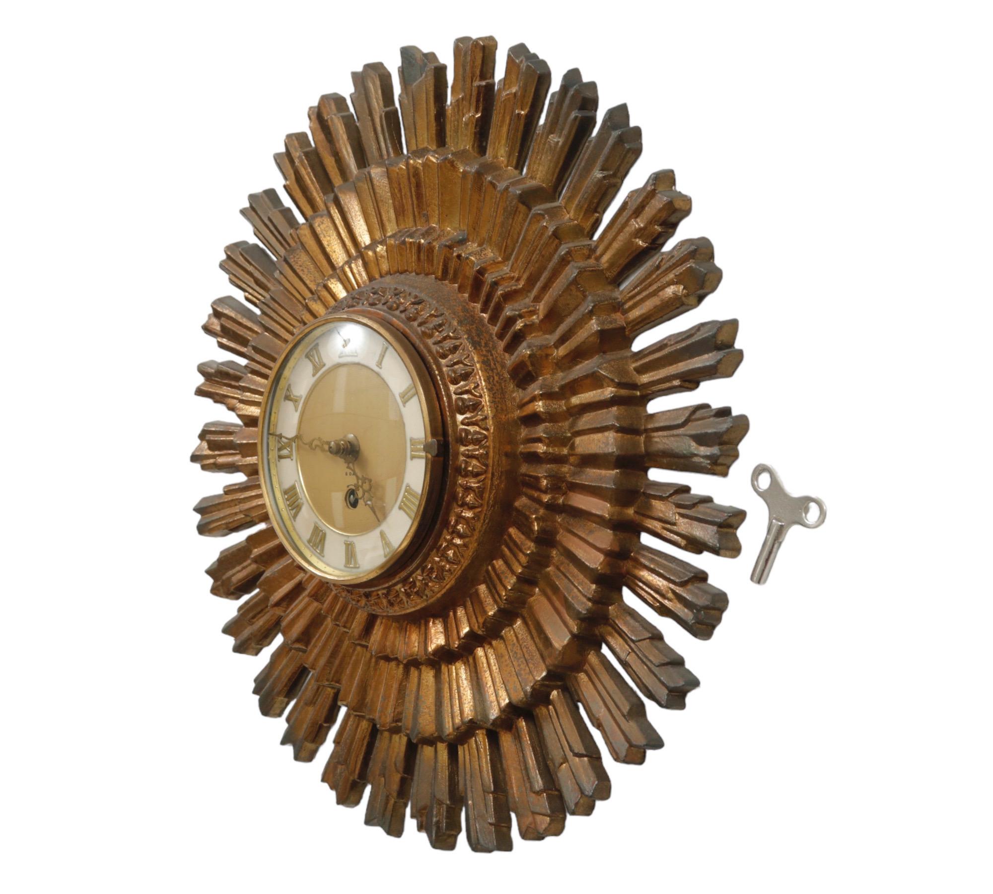 A round, decorative Hollywood Regency wall clock, circa 1960. A large metal sunburst frame in the original finish surrounds a clock face with a glass front that opens. Roman numerals and ornate cast brass hands tell the time. The mechanism winds