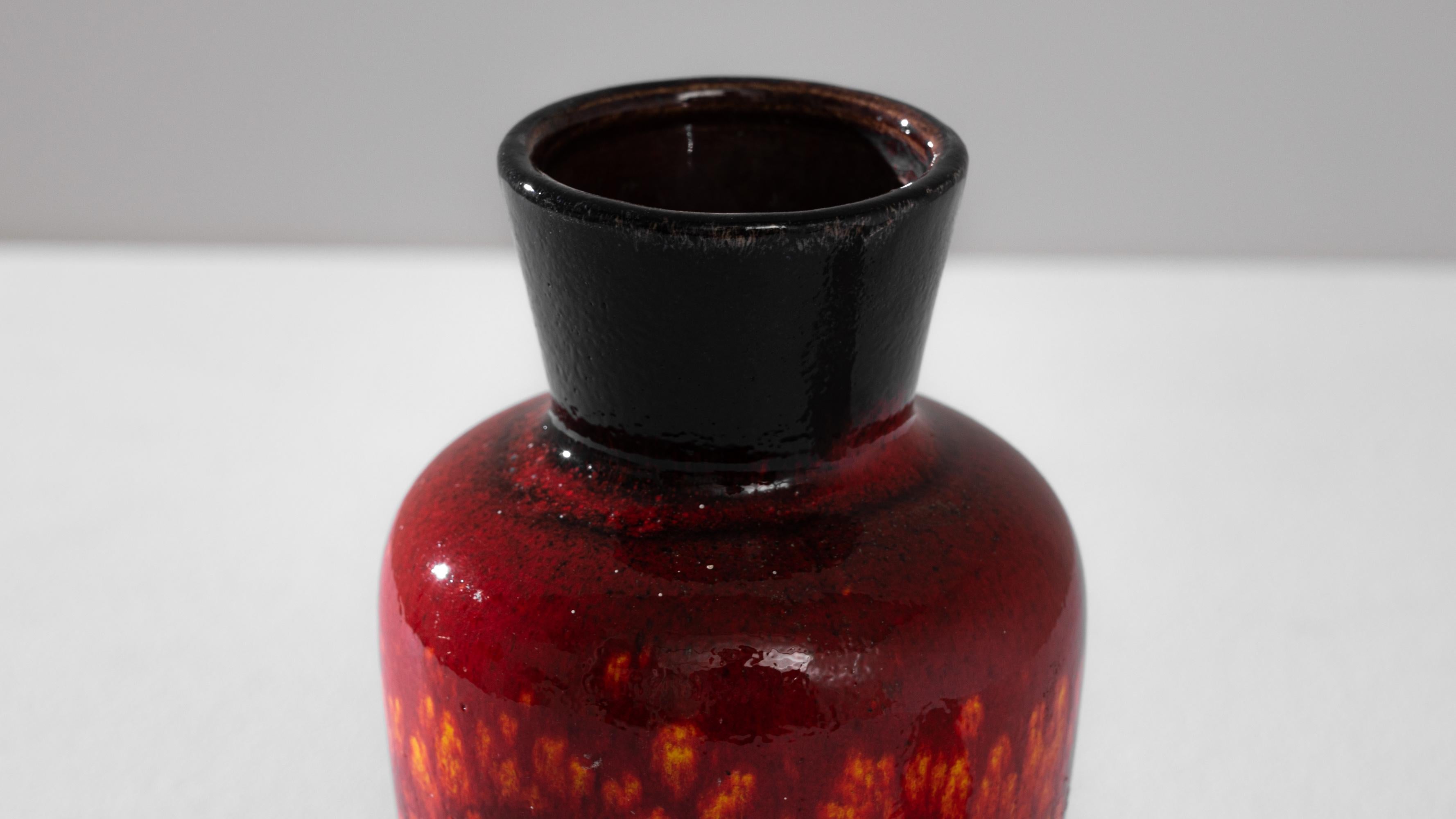 This striking 1960s German ceramic vase is a vibrant testament to the era's bold aesthetic and design ingenuity. The deep, fiery red glaze, speckled with intense bursts of yellow and black, makes a dramatic statement, while the sleek, minimalist
