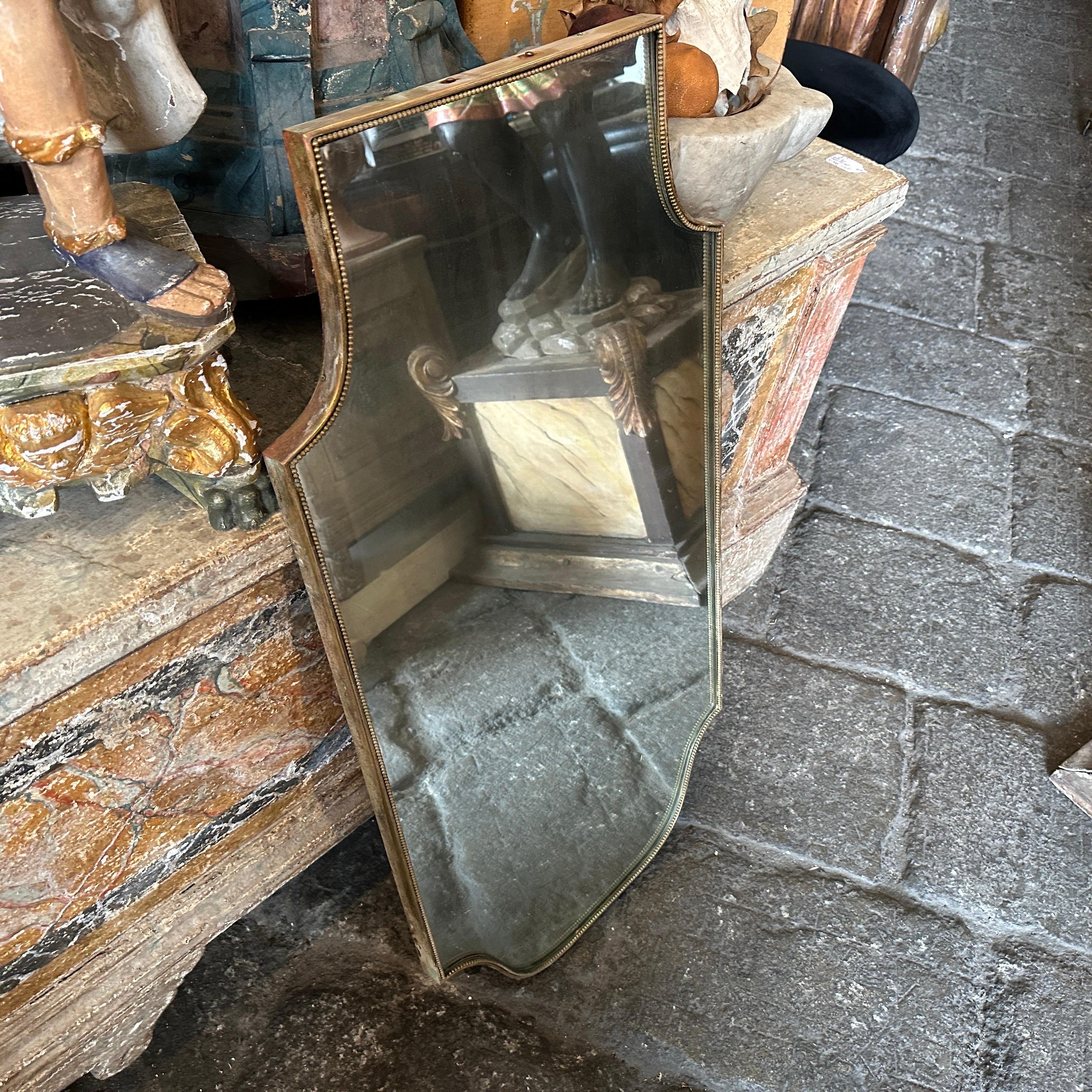 Specchio da parete in ottone a forma di scudo, disegnato e prodotto in Italia negli anni Sessanta nello stile di Giò Ponti; è in buone condizioni, l'ottone presenta una patina originale con segni d'uso e d'età; l'ottone è un materiale che può essere