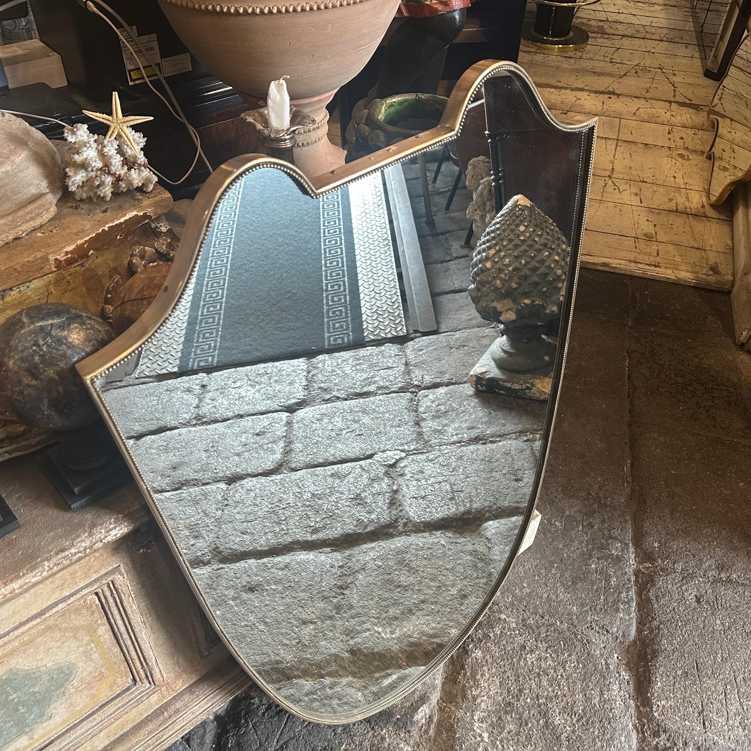 A big brass shield-shaped wall mirror, designed and manufactured in Italy by Luigi Fortuna in the style of Gio Ponti. The solid brass frame is in its original patina with signs of use and age, while the mirror itself is in perfect condition. This