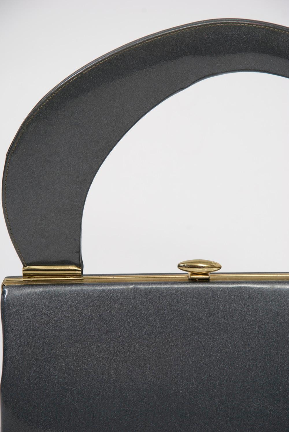 1960s large handbag featuring an unusual sweep of a handle, lending it a modernist aesthetic, and crafted in a most desirable color and material - gunmetal patent. Simple brass frame and closure keeps it streamlined. The interior is of beige fabric