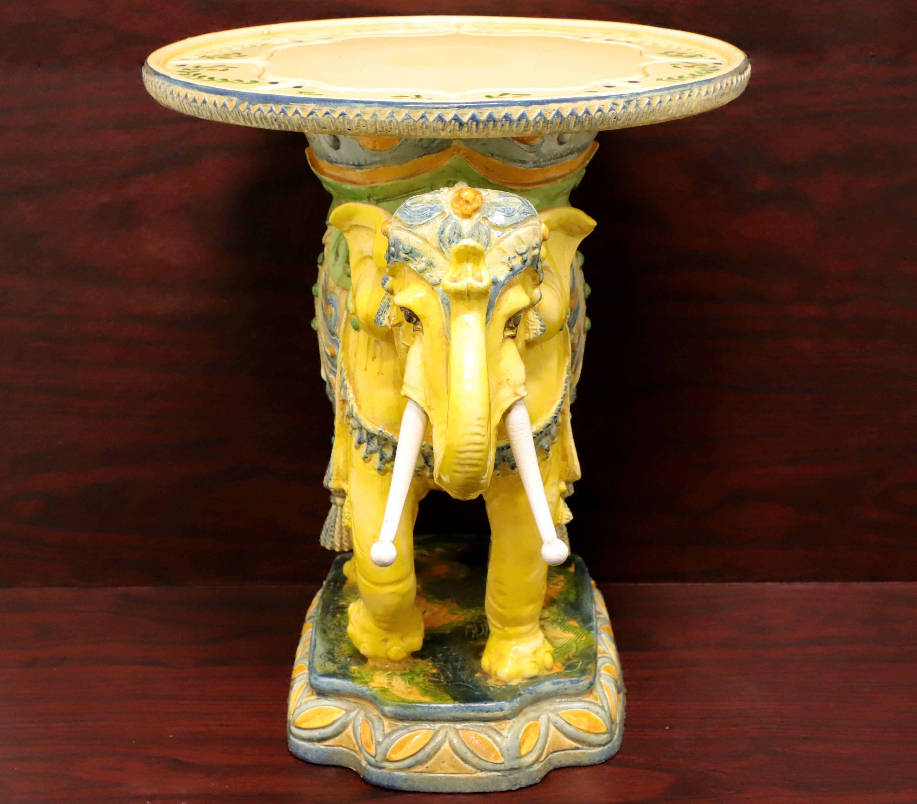 A ceramic plant stand in the Hollywood Regency style depicting an elegantly dressed elephant with a round flat surface top ideal for a plant. Unsigned, artist unknown. Multi-colored, mostly shades of yellow, blue, green & orange, ceramic, glazed and