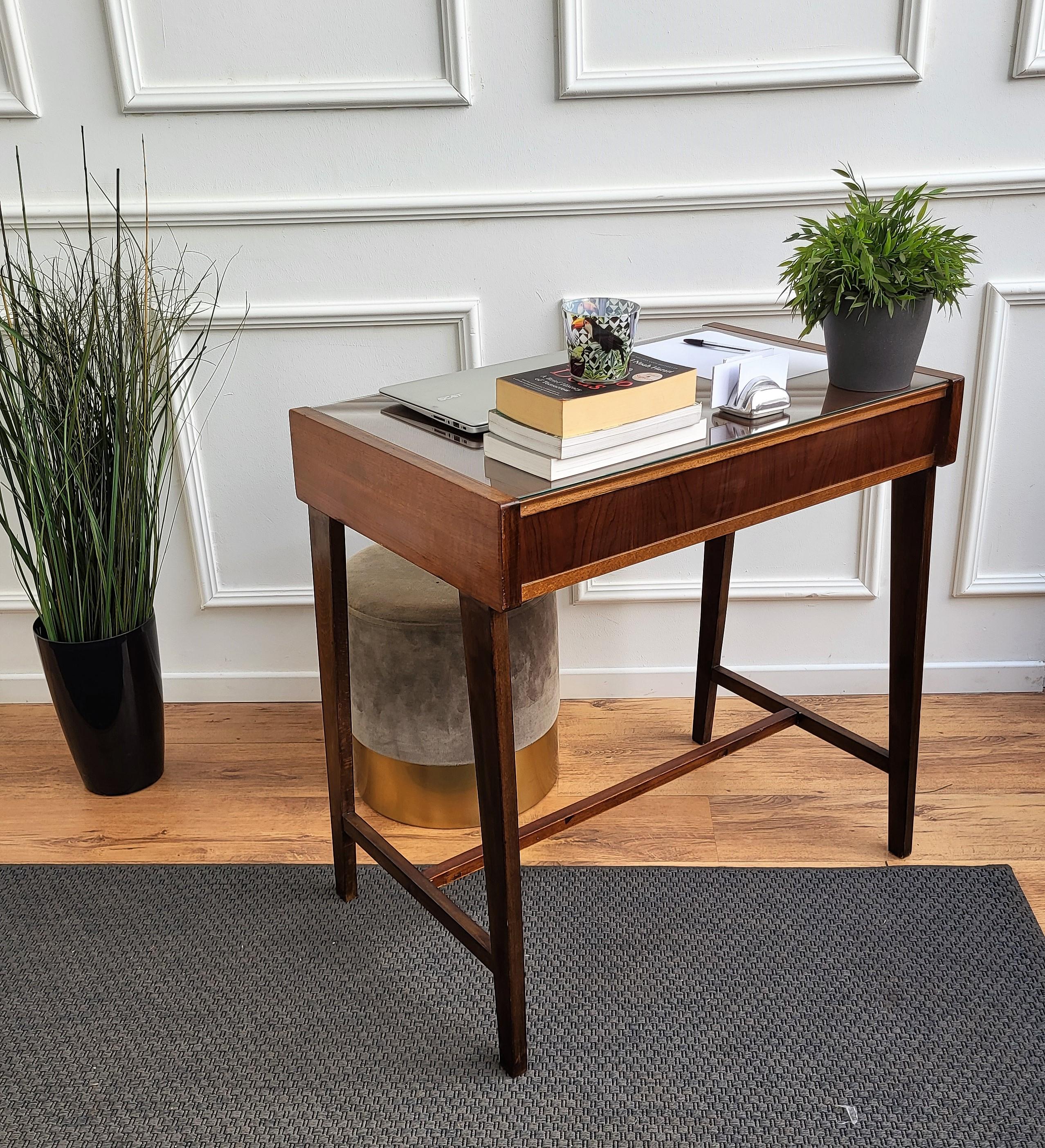 Very elegant Italian Mid-Century Modern small desk or writing table with stylish legs and veneer wood with a central drawer. This beautiful piece is completed by the glass top and the brass drawer handles. Ideal in any room as a small stand alone