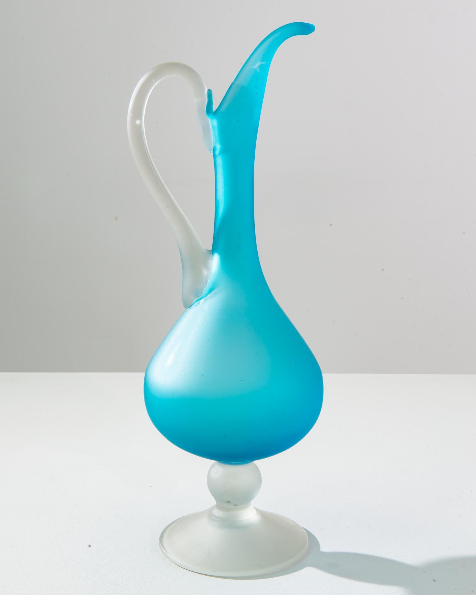 Immerse yourself in the cool, serene beauty of this 1960s Italian blue glass jug, a stunning piece that reflects the free-spirited creativity of the era. The jug’s body boasts a captivating sky blue color that seems to hold the essence of the