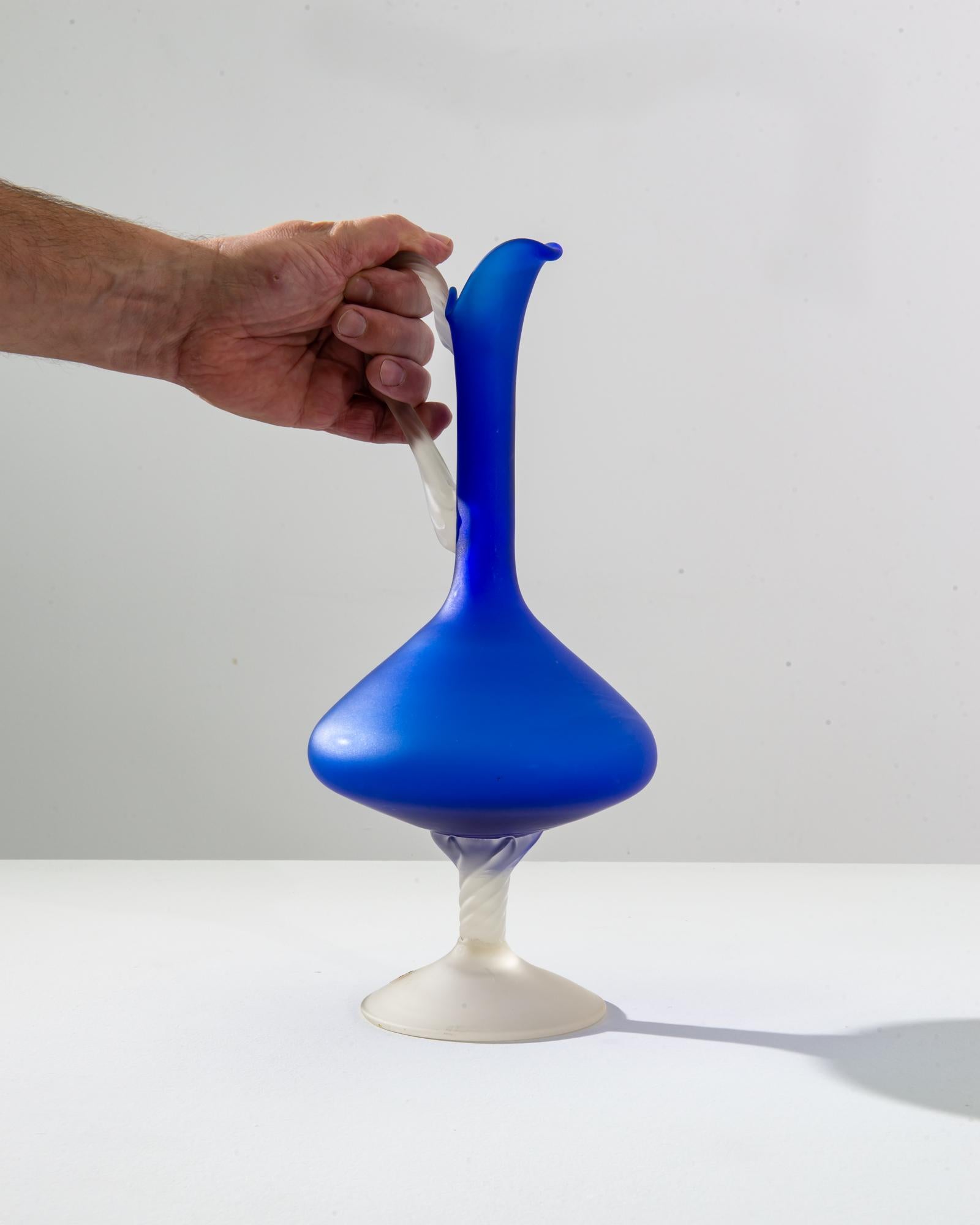 This 1960s Italian blue glass jug is a quintessential piece of mid-century design, its rich cobalt hue reminiscent of the deep waters of the Mediterranean. The jug's organic, bulbous base flows into a slender, flared neck, culminating in an