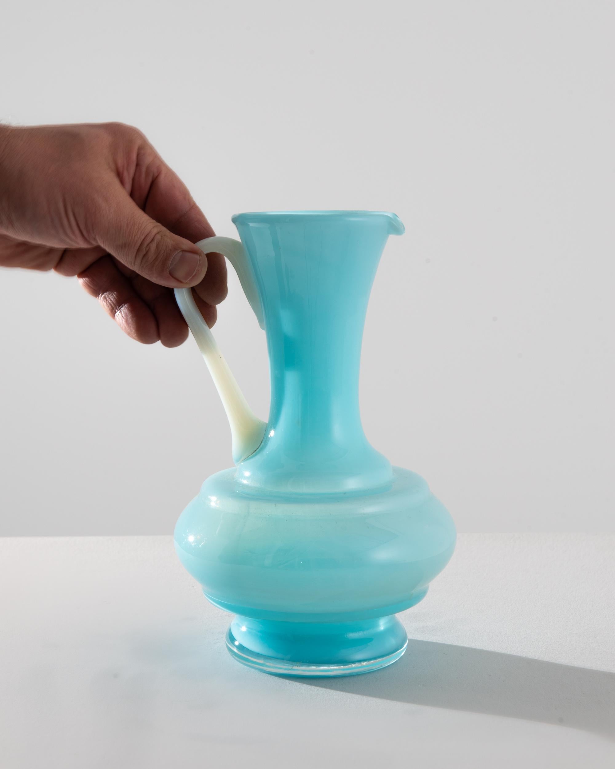 Introducing the captivating 1960s Italian Blue Glass Jug, a masterpiece of vintage charm and elegance. This exquisite piece is a symphony of pastel blue and creamy white, evoking the carefree spirit of a bygone era. The bulbous base, crafted with a