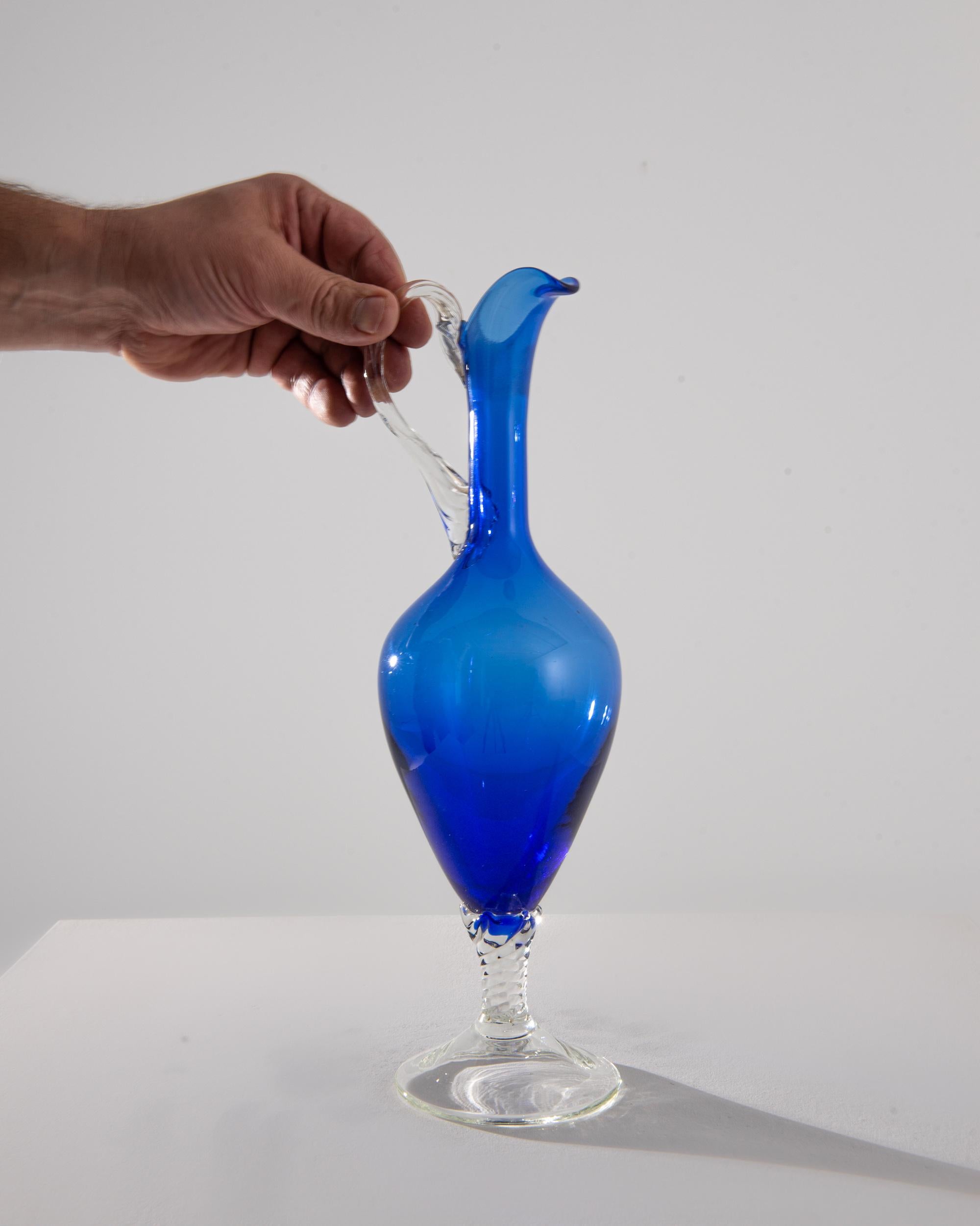 This exquisite 1960s Italian Blue Glass Jug, with its vivid cobalt blue hue and elegant clear glass handle, is a testament to the era's innovative design and glassblowing artistry. Its alluring shape is reminiscent of an elongated teardrop,