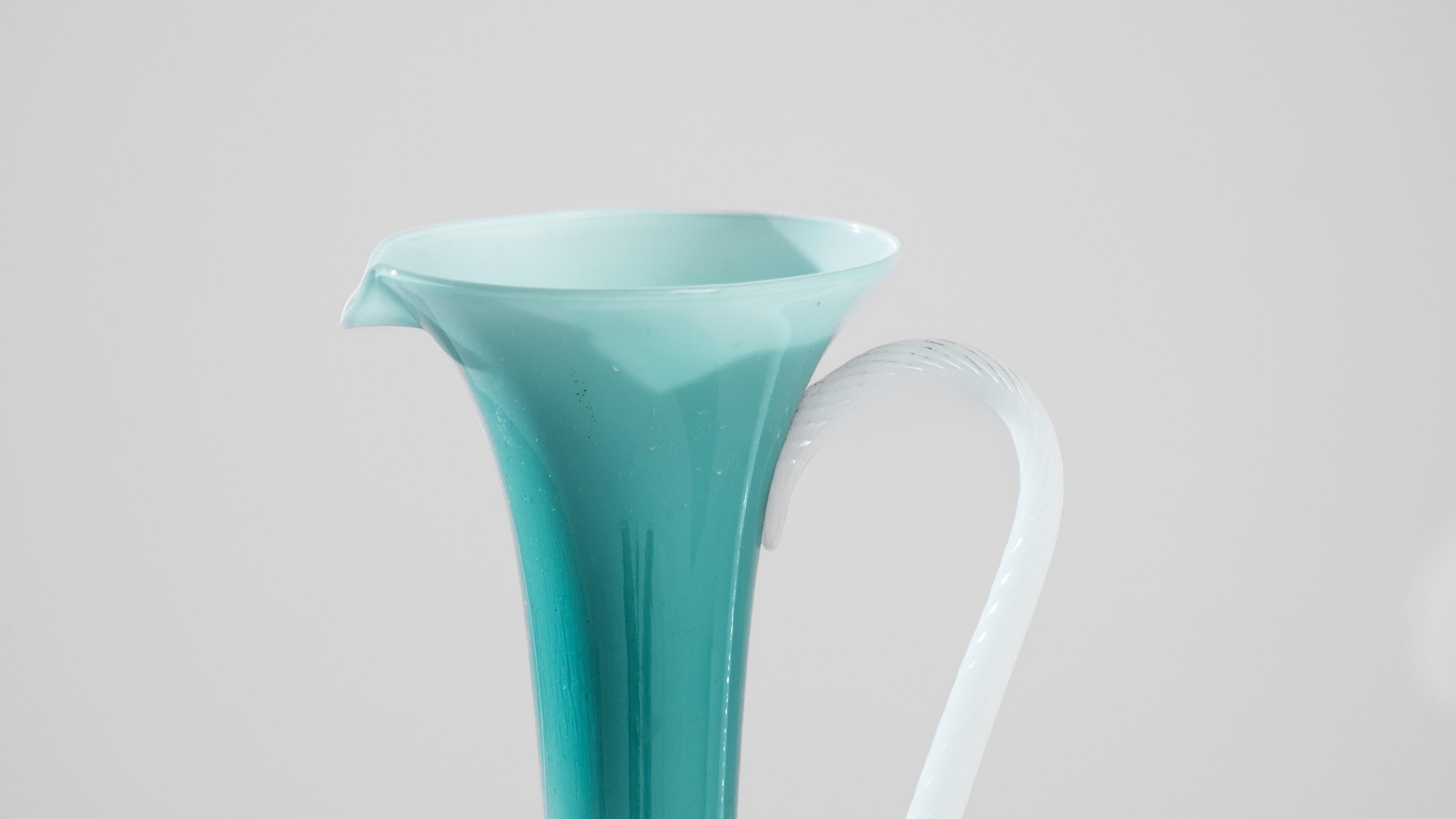 This 1960s Italian green glass jug is a stunning example of vintage elegance and timeless design. Crafted during a period where attention to detail and form was paramount, the jug exudes a delicate balance between beauty and function. Its slender