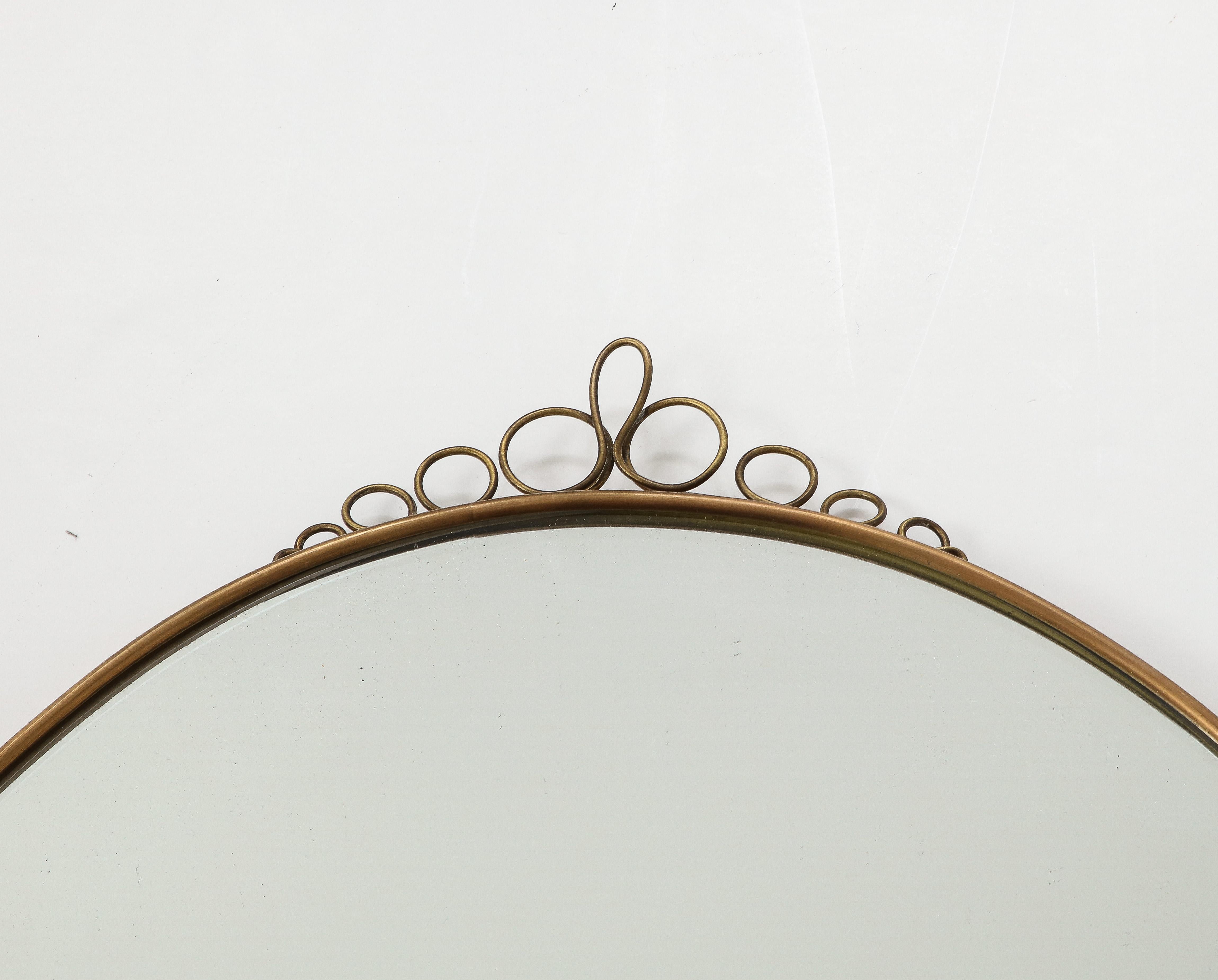 1960s Italian modernist elegant oval brass wall mirror with decorative brass scroll elements on central top and bottom frame. This lovely mirror has a beautiful patina on the brass and illustrates in its playful scroll decorations the Italian