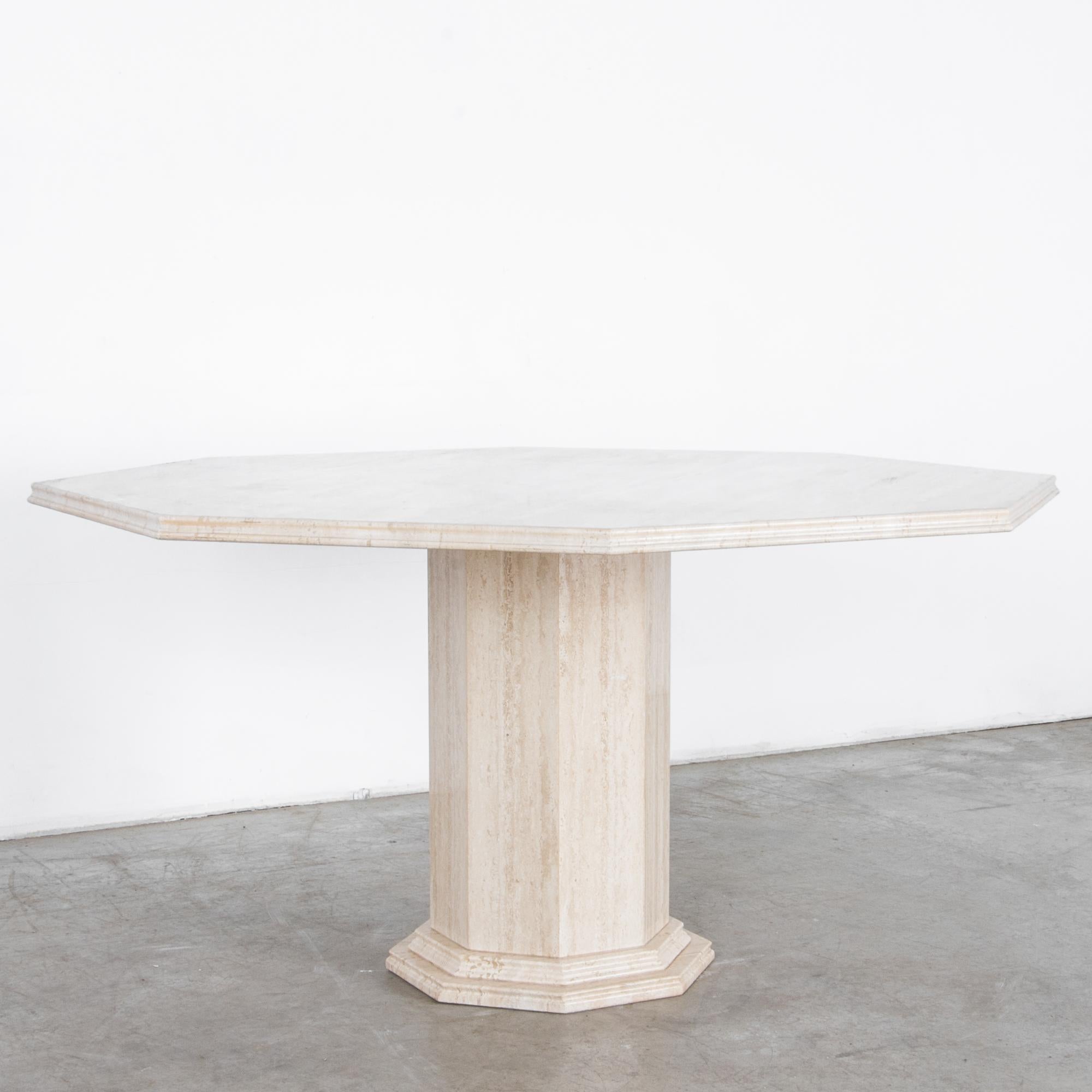 A cantilevered octagonal slab of light travertine rests on a faceted round pedestal, in this stylish and simple dining table. The tabletop is carved with an ogee edge profile, and framed below by a profiled column base, in a neoclassical inflected