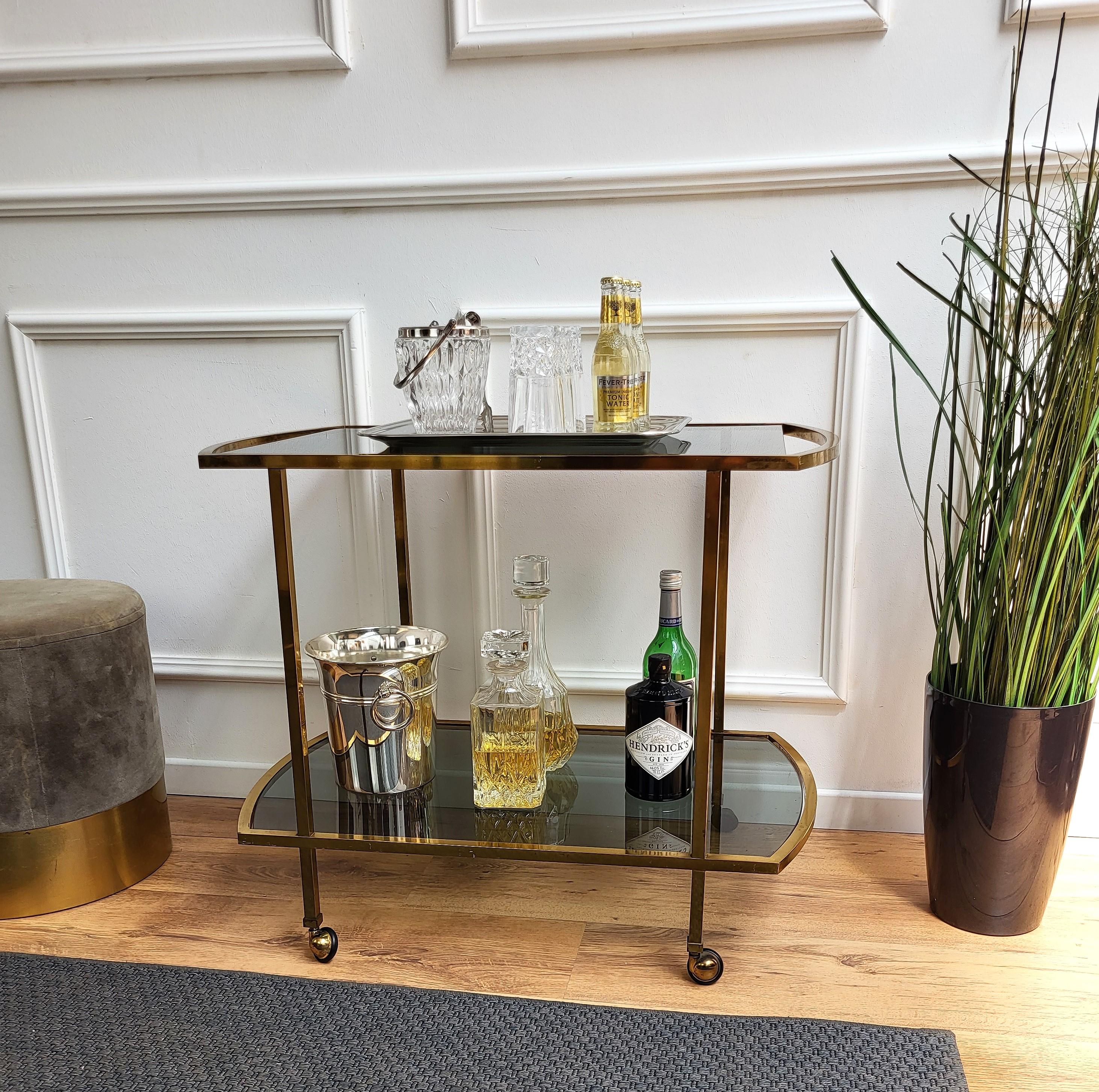 Beautiful and stylish 1960s Hollywood Regency Italian two-tier brass and smoked glass bar cart. A great piece that perfectly adds to every home decor the typical glitz, glamour and gold of Hollywood Regency style, with a nod to Art Deco decadence