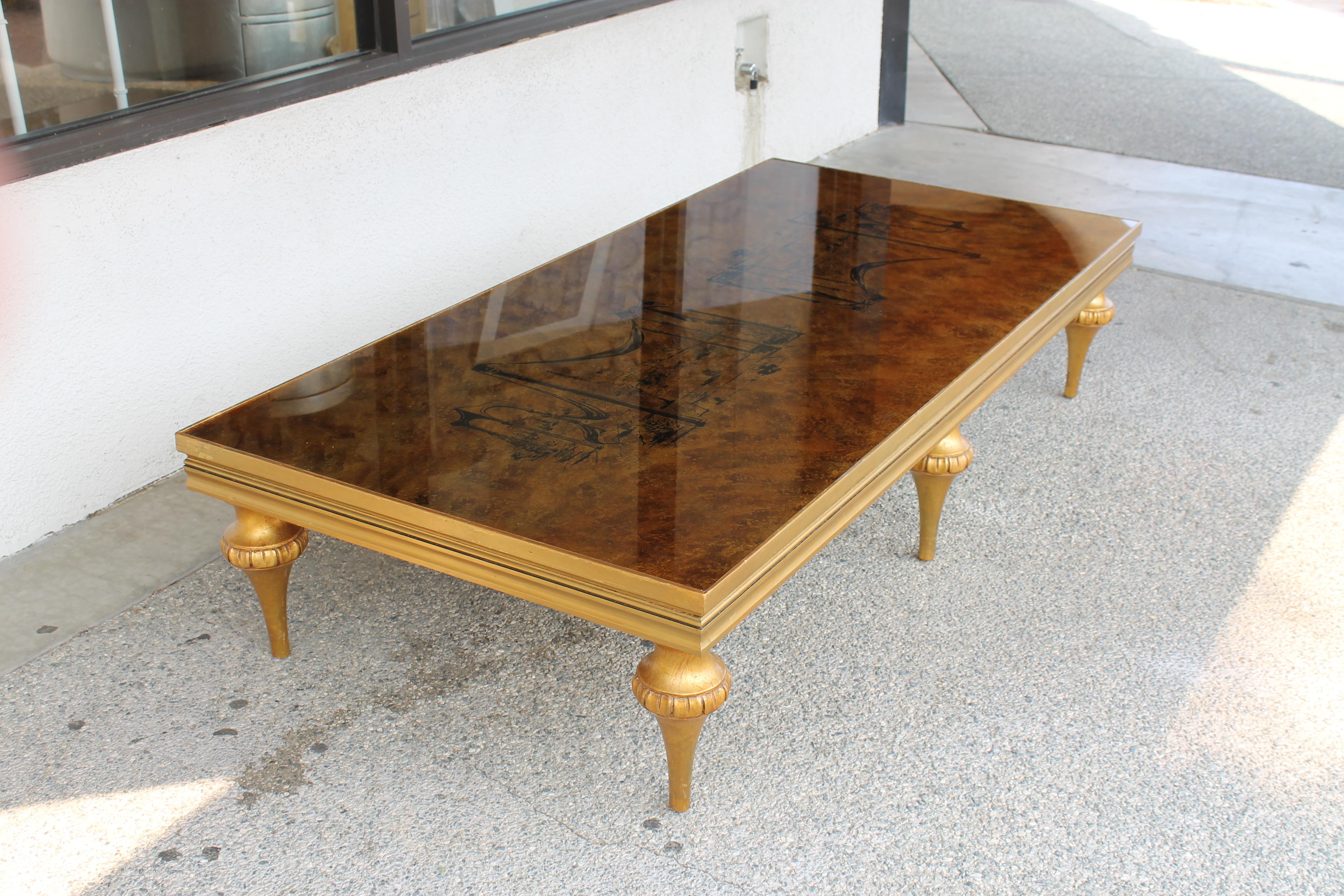 A huge (6 foot) Venetian style coffee table with reverse painted glass top surface, rich gold with black surrealist landscape. Carved gilt painted wooden legs. Table measures 72