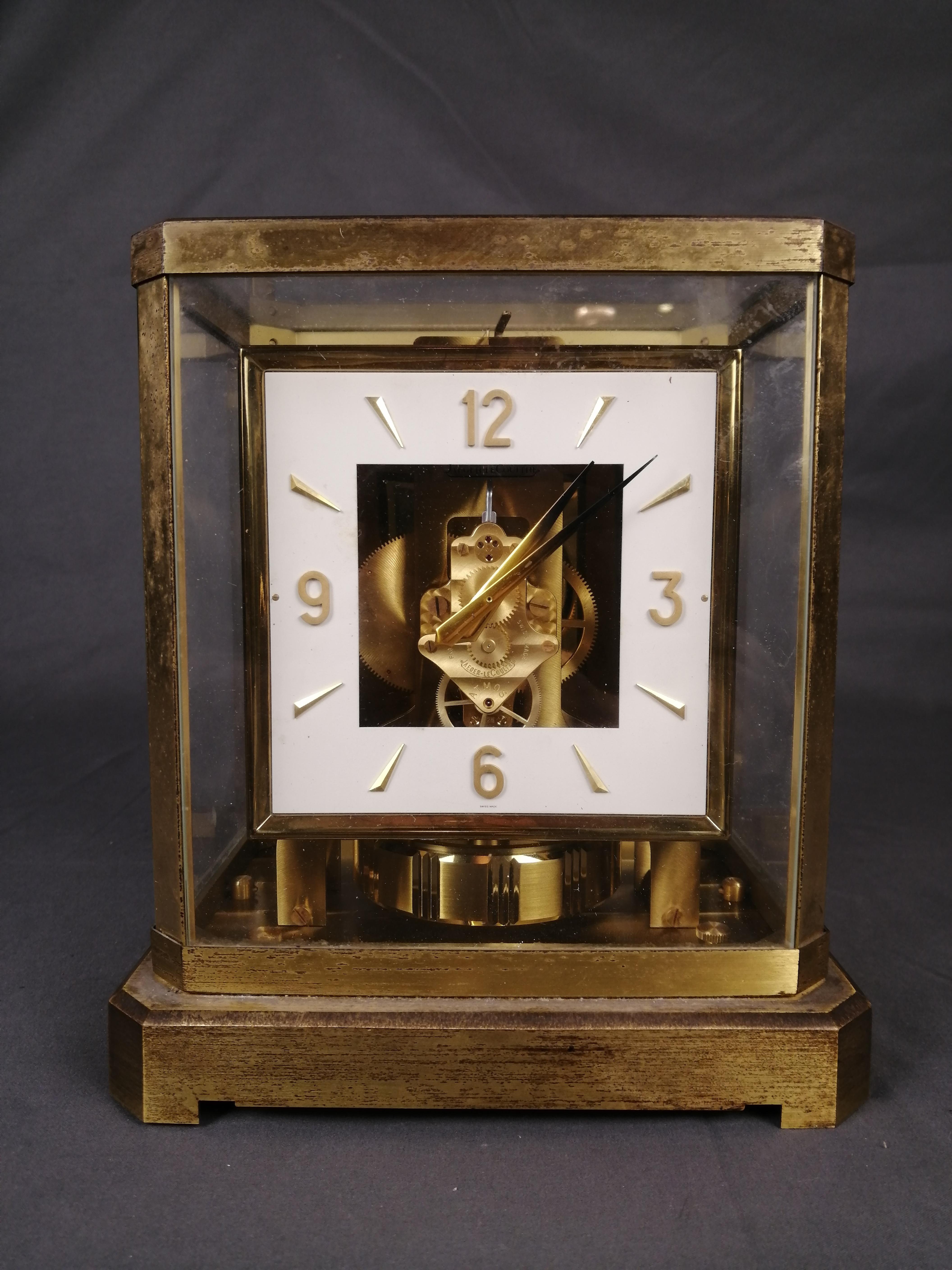 A 1960's Swiss Atmos mechanism clock by Jaeger-LeCoultre. Serial number 134555. The squared dial with white face has prisms and numbers 3, 6, 9 and 12. The hands are sword-shaped. The brass base and showcase with glass shows wear. 

