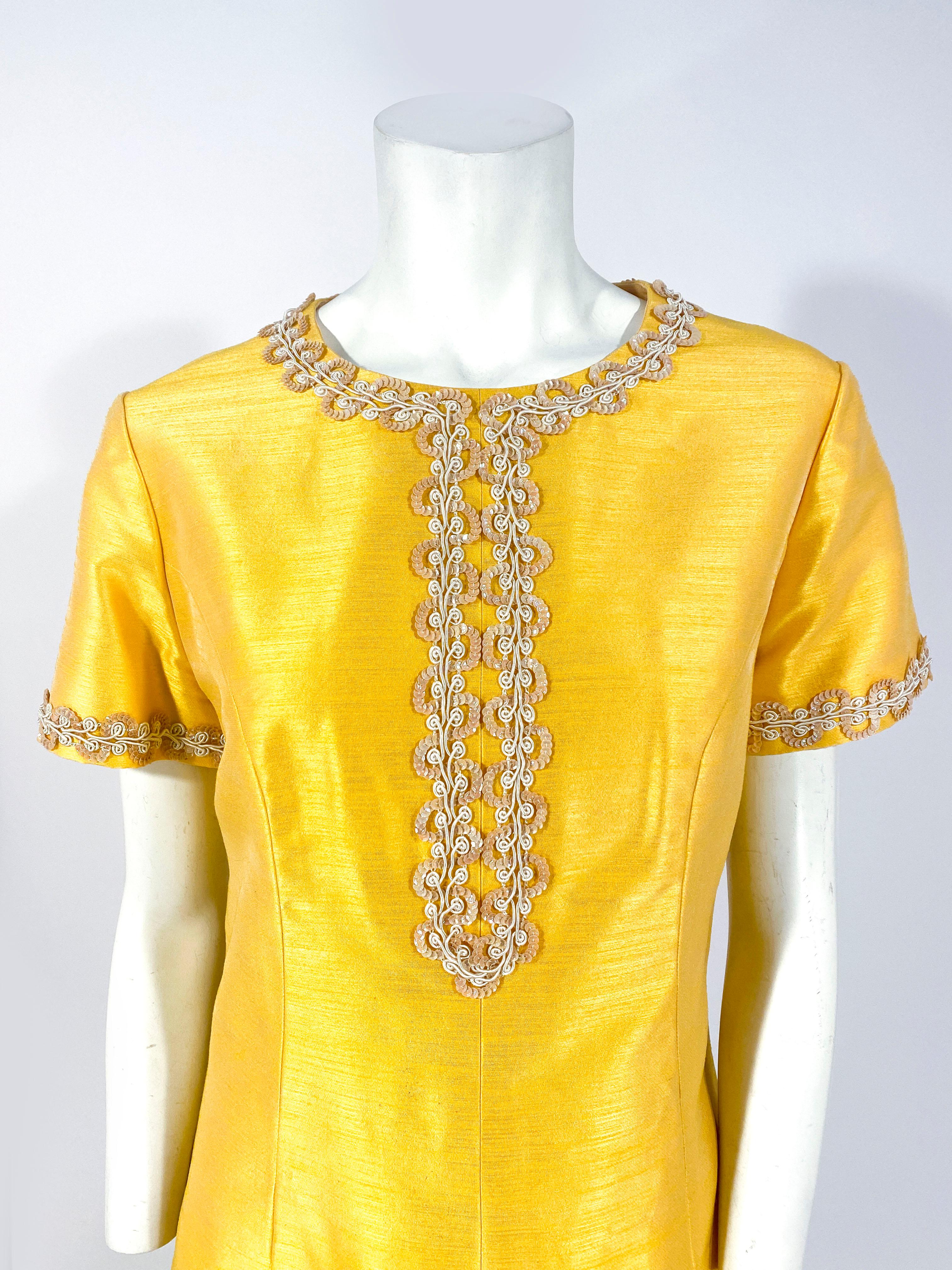 1960s Joseph Magnin Gold/yellow A-line dress with short sleeves and sequin/cord trim along the neckline, down the center, and on the sleeves. There are enlarged post darts to provide shape to the body of the dress and the back has the metal zipper