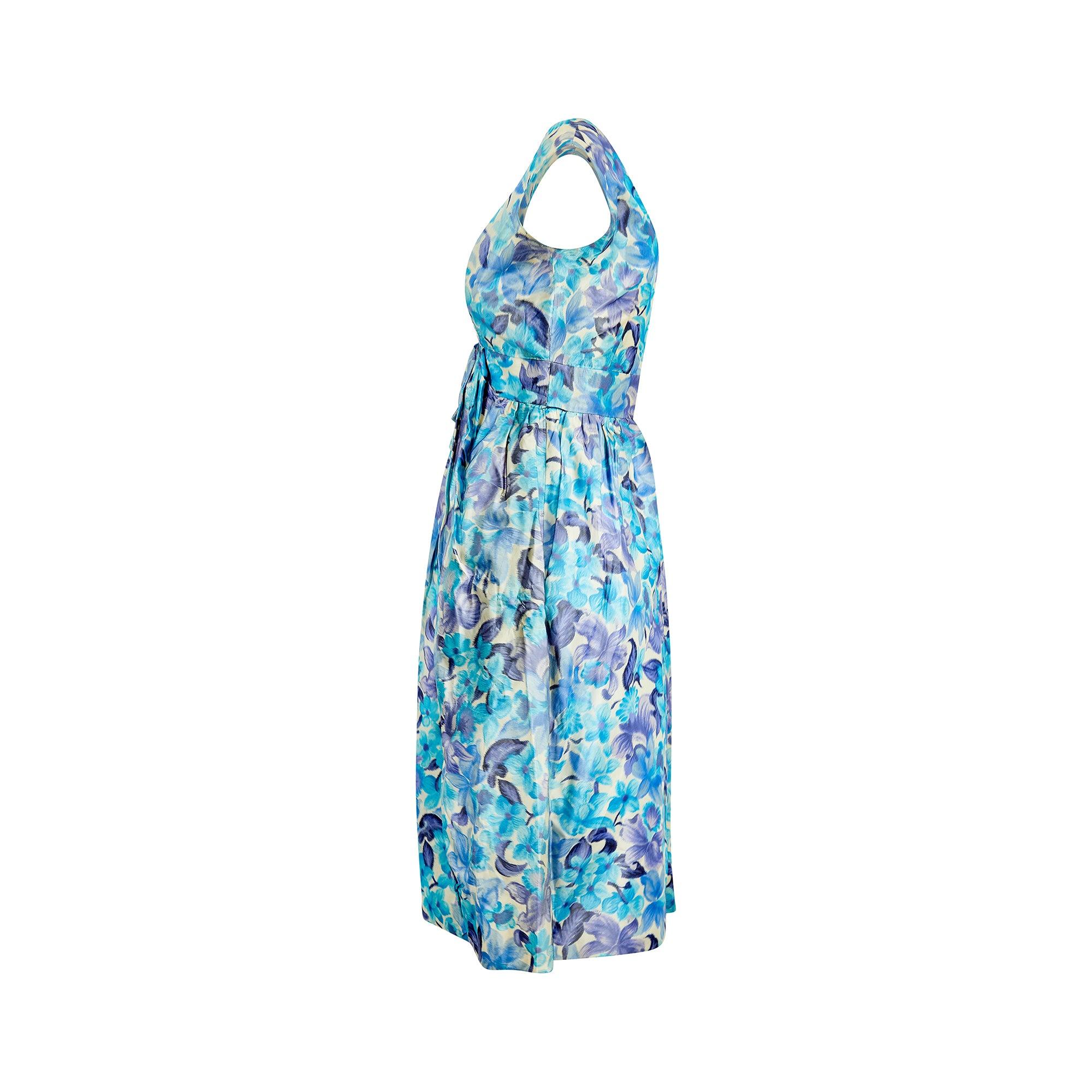 Dieses Kleid von Koupy Couture ist aus Seide gefertigt, die mit leuchtend blauen und fliederfarbenen Blumen bedruckt ist. Der weite V-Ausschnitt, ein feminines Merkmal vieler Designs der 1960er Jahre, lässt die Schlüsselbeine und das Dekolleté zur