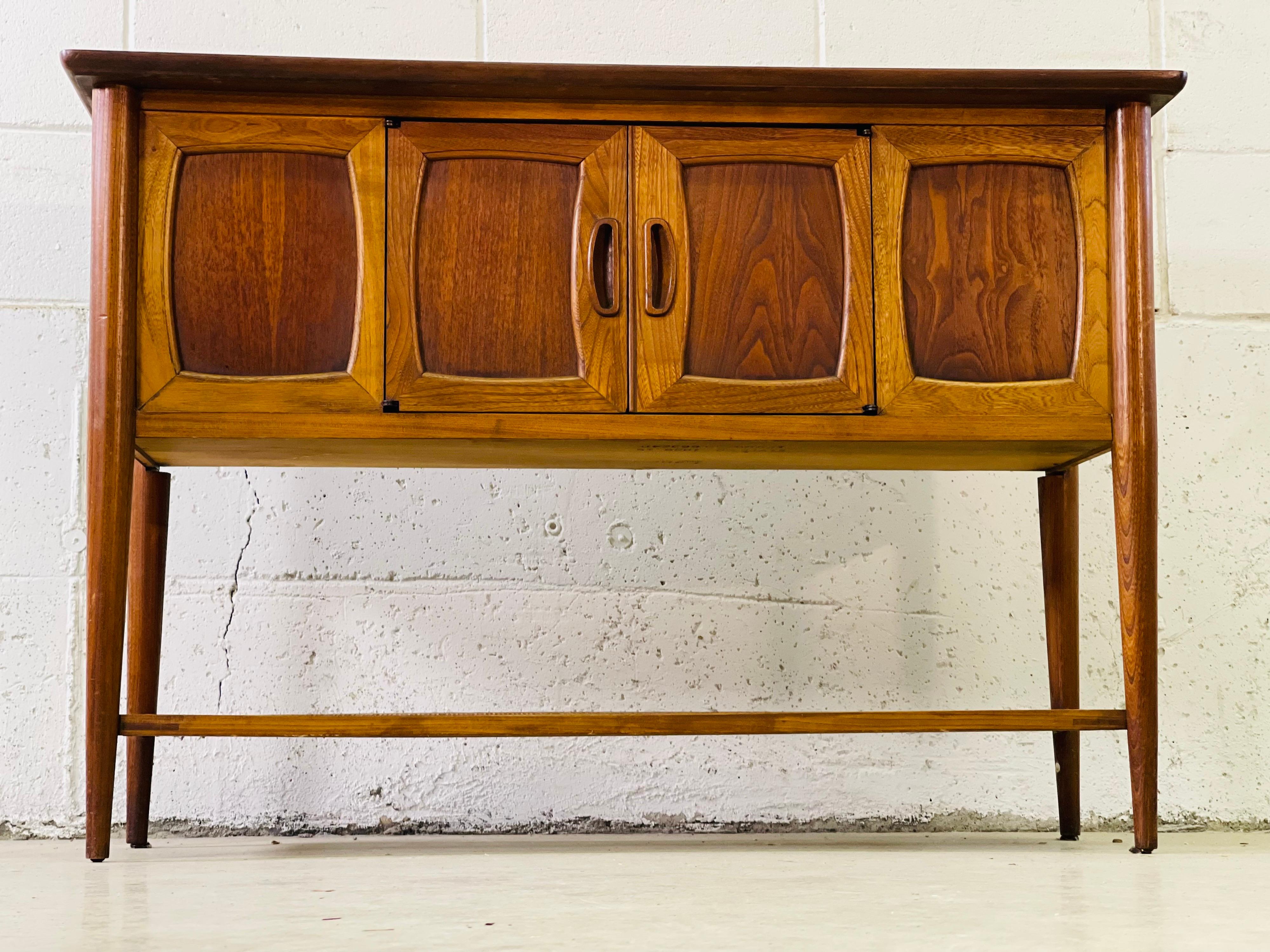 1960s walnut console table by Lane Furniture Company. The table has two doors in the front for open storage inside and a rattan shelf on the bottom. The top of the table is faux wood laminate. Marked underneath.
