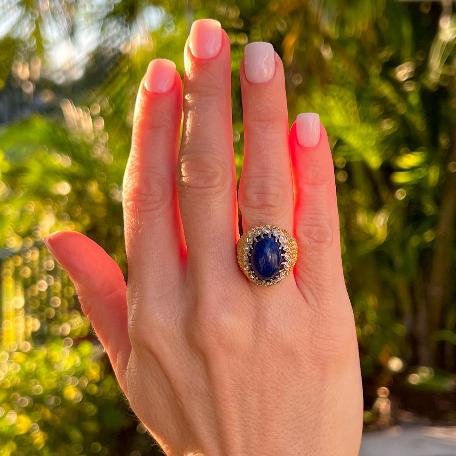 Beautiful lapis lazuli cocktail ring handcrafted in textured 18 karat yellow gold. The ring features a cabochon blue lapis gemstone set with 17 surrounding diamonds weighing approximately .15 CTW. The ring measures 20mm in width, and is currently