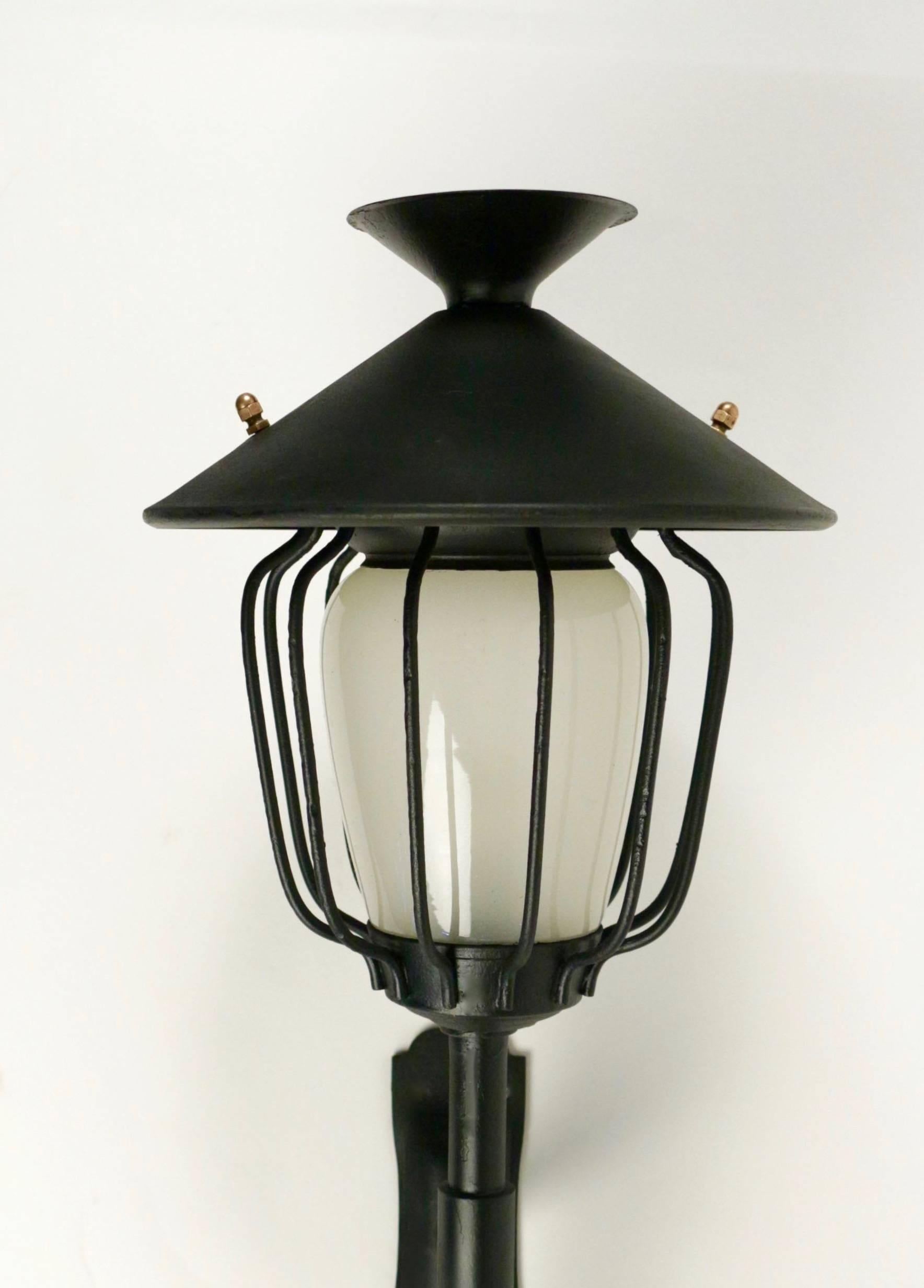 Large pair of Lanterns wall lights from Maison Honoré from the 1960s.

Composed of a long spindle-shaped arm on which rests an original satin-glass lantern, enclosed in an openwork cage by black wrought iron rods and dressed in a 