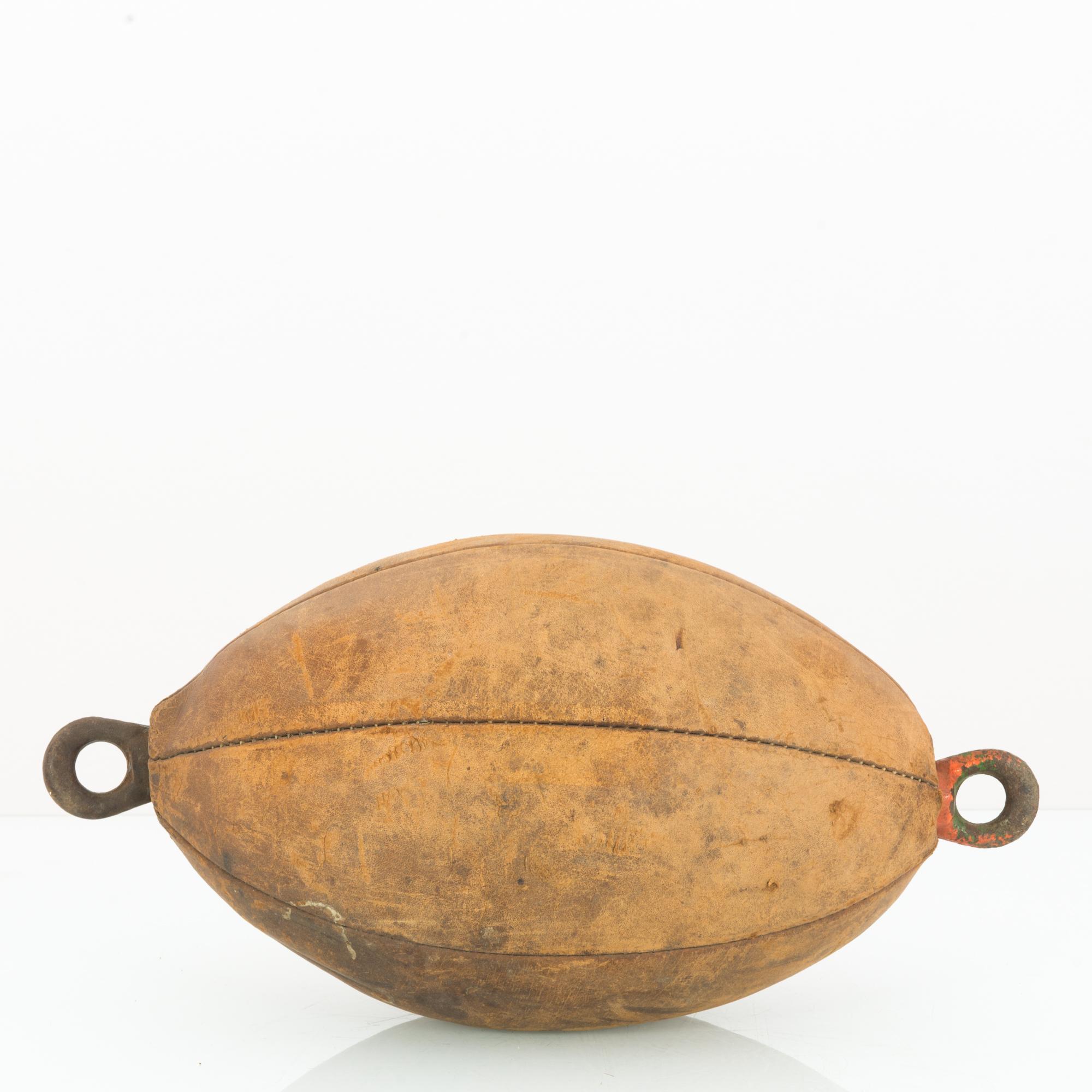 A leather boxing ball from Czechia circa 1960. A lozenge of tan leather filled with padding. Metal rings at each end allow the ball to be hung as a target for striking practice. The weathering of the leather and the oxidation of the metal rings have