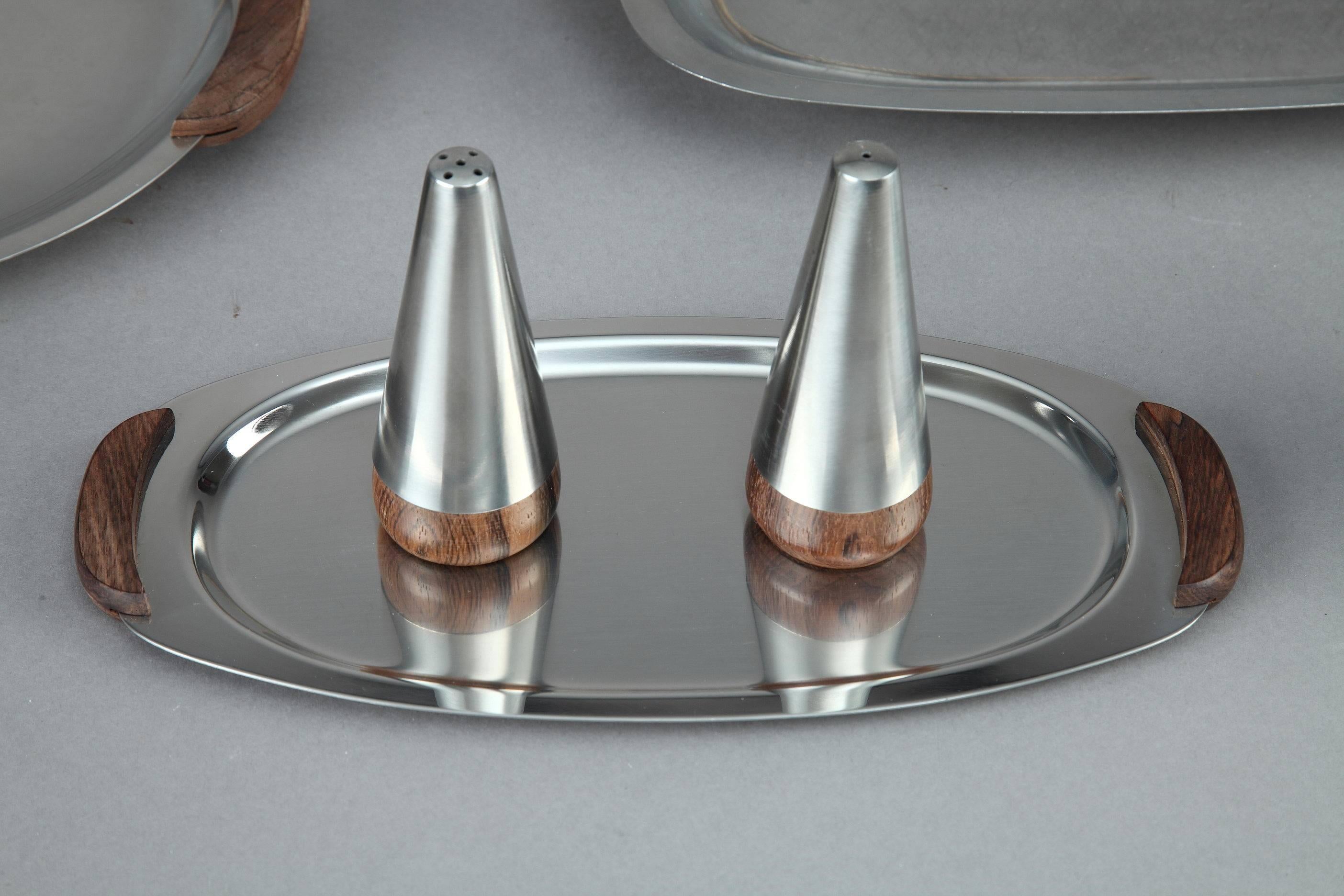 Stainless steel table service set from the 1960's. It is composed of: 3 deep platters, 3 smaller, shallow platters, 4 candlesticks, 3 pots (2 sauce, 1 milk), Salt and pepper shakers. The platters (two of which have covers) have wood handles. Three