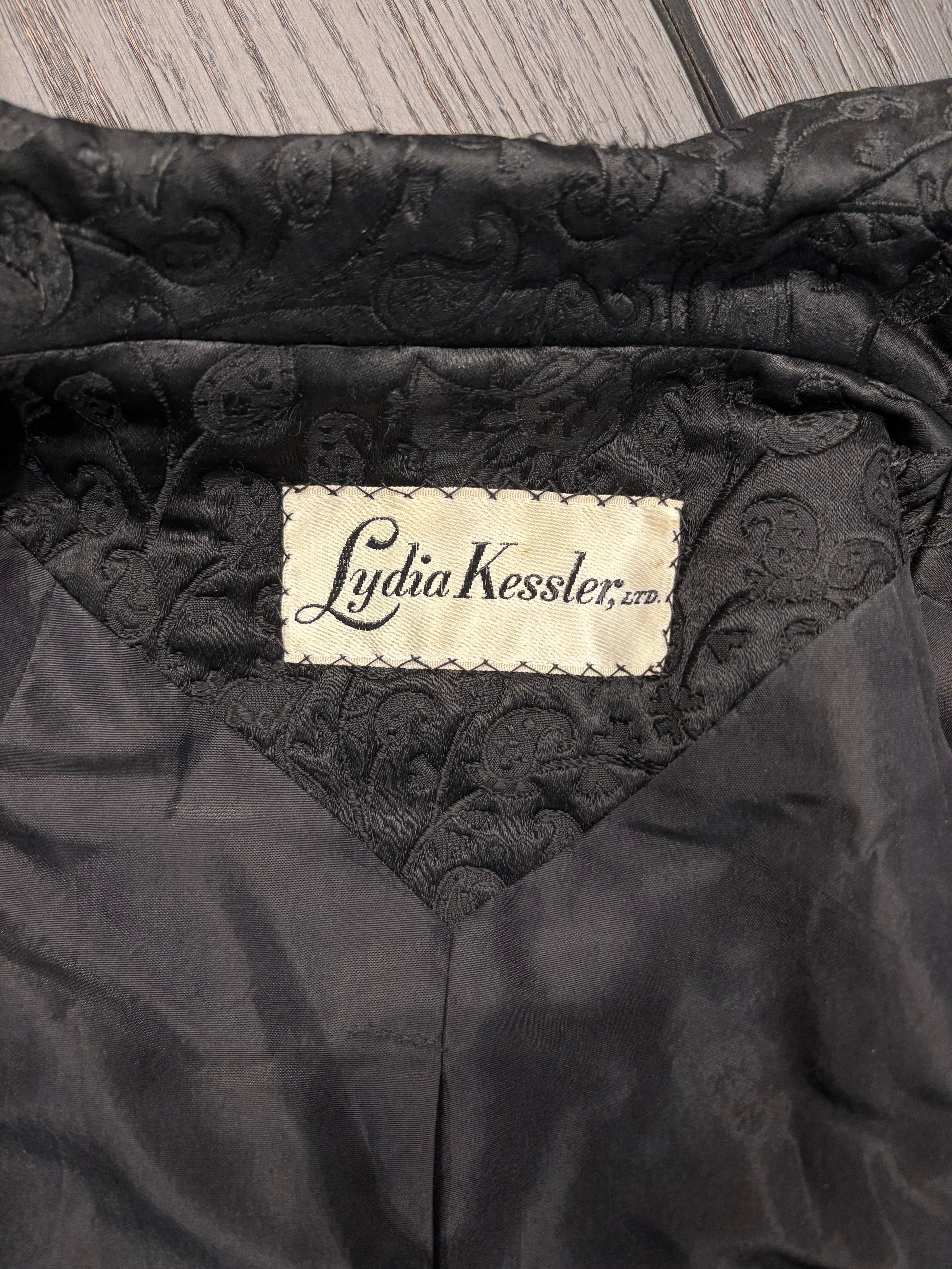 Un extraordinario abrigo princesa de brocado negro de Lydia Kessler Ltd. de los años 60, impecablemente confeccionado en un tejido jacquard paisley de rica textura que capta la luz con sutil brillo. Este abrigo de noche de cuerpo entero evoca la